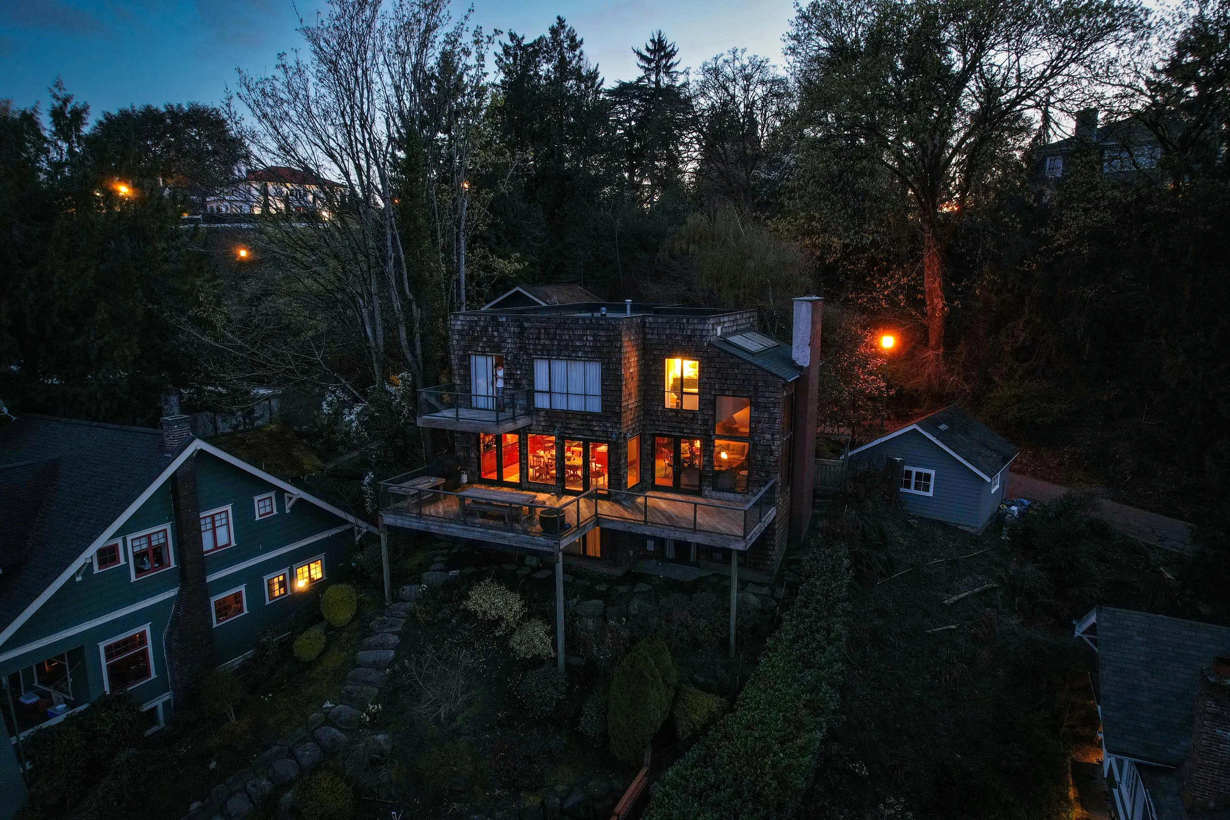 Aerial view of a modern wooden house with large glass windows and multiple decks, illuminated from within, surrounded by trees at dusk.