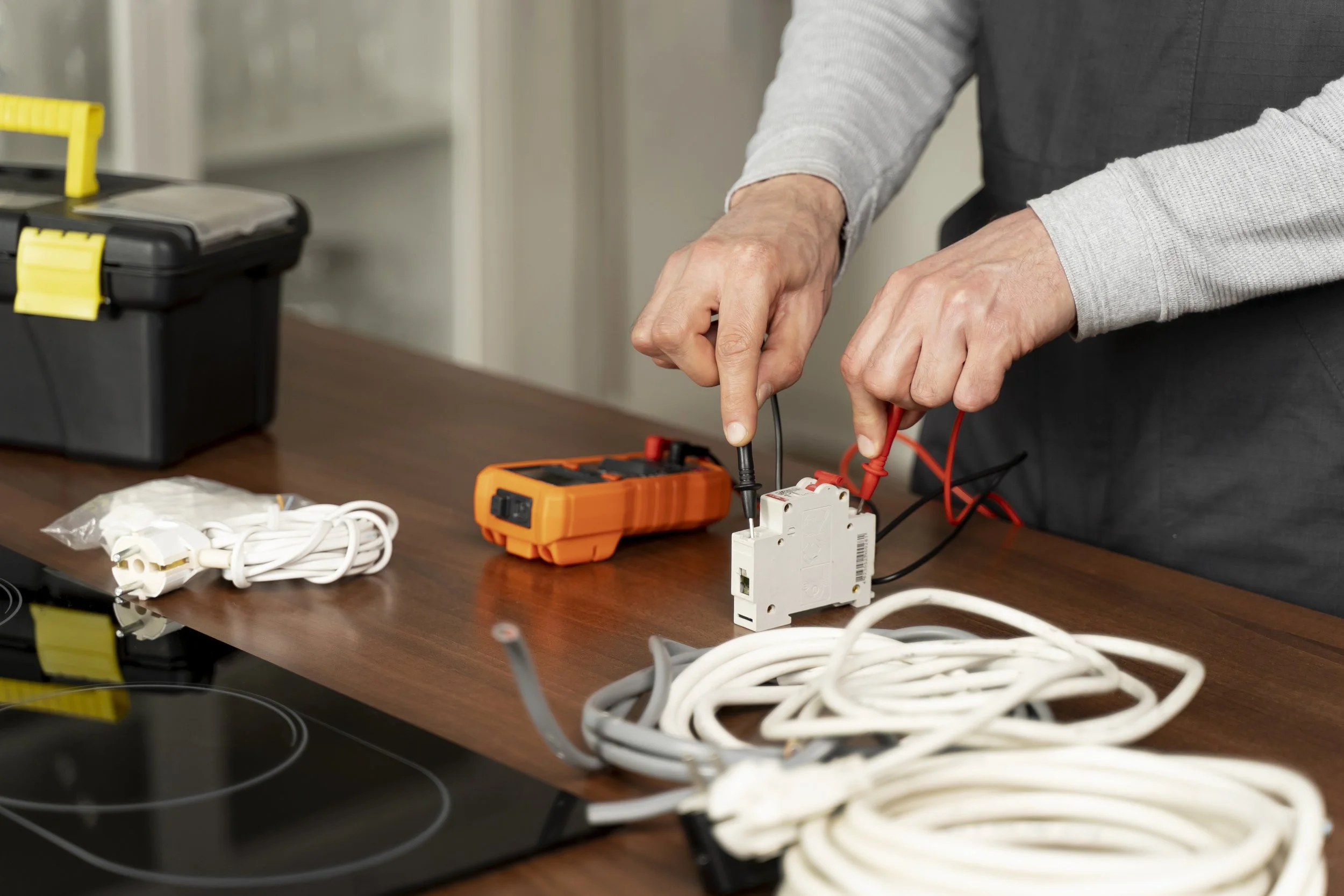 Person working with electrical tools, using a multimeter on a circuit breaker, with various cords and a toolbox on a wooden table.