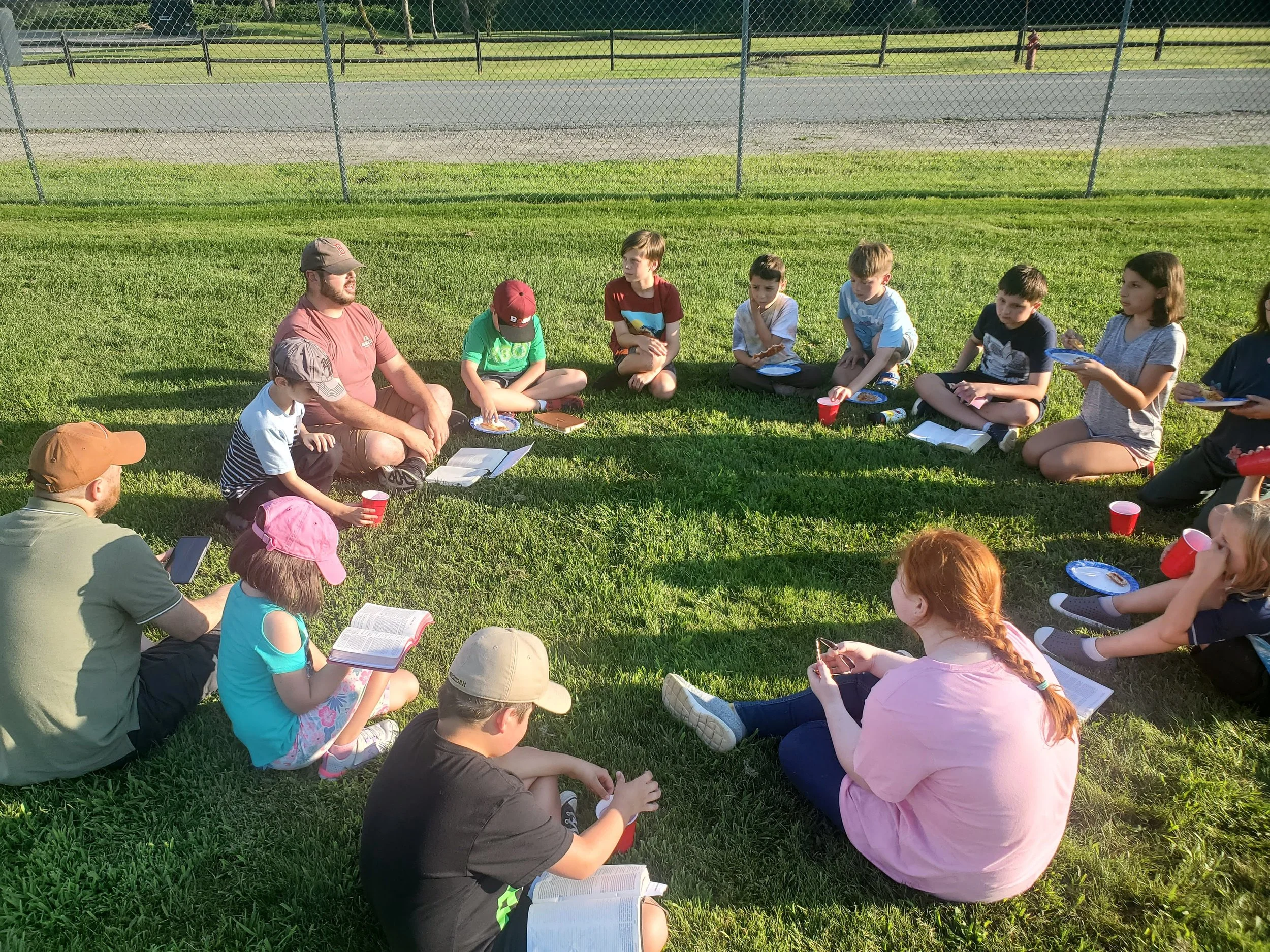 A group of children and adults sitting in a circle on the grass outdoors, with some holding books, notebooks, or drinks. They appear to be participating in a group activity or discussion. There is a chain-link fence and a paved path in the background.