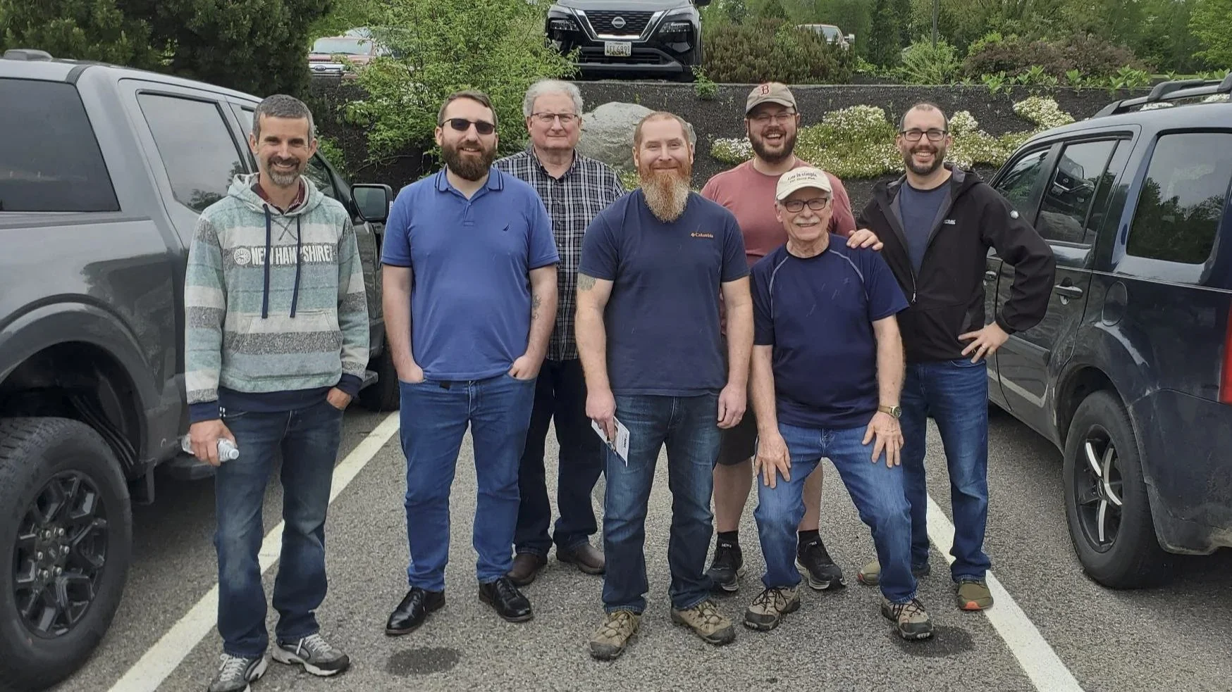Group of nine men standing in a parking lot, surrounded by cars and greenery, smiling at the camera.