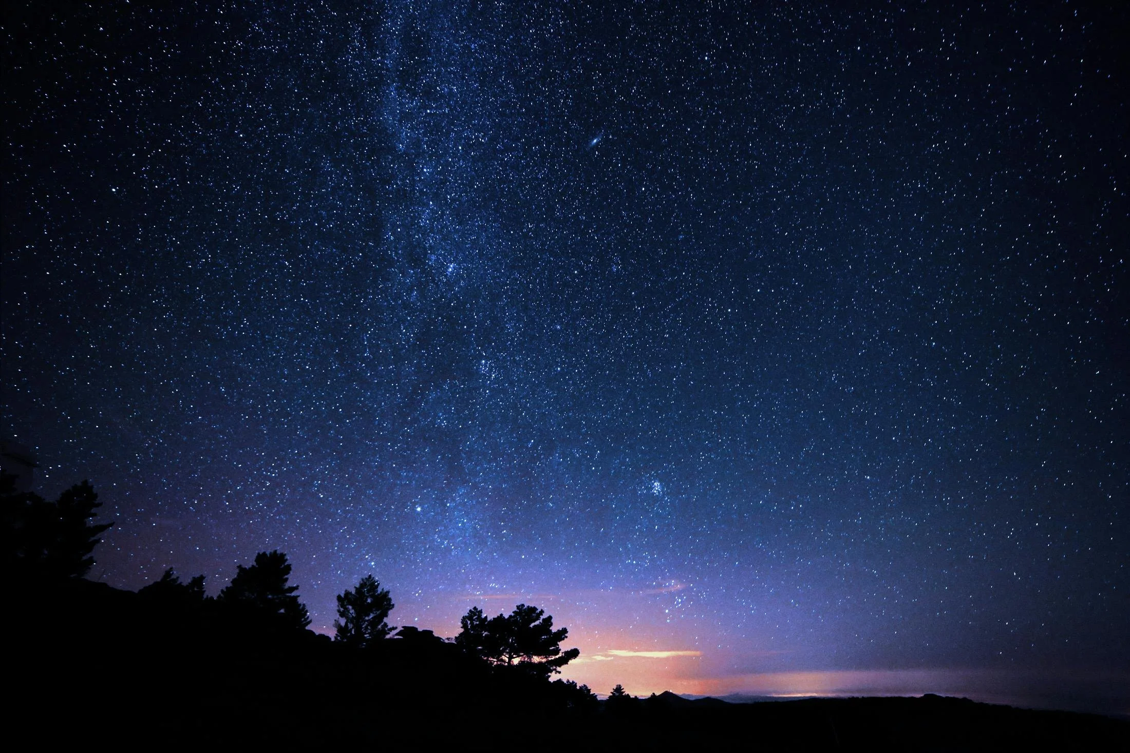 Starry night sky with many stars and a faint Milky Way, with silhouette of trees and mountains at the horizon.