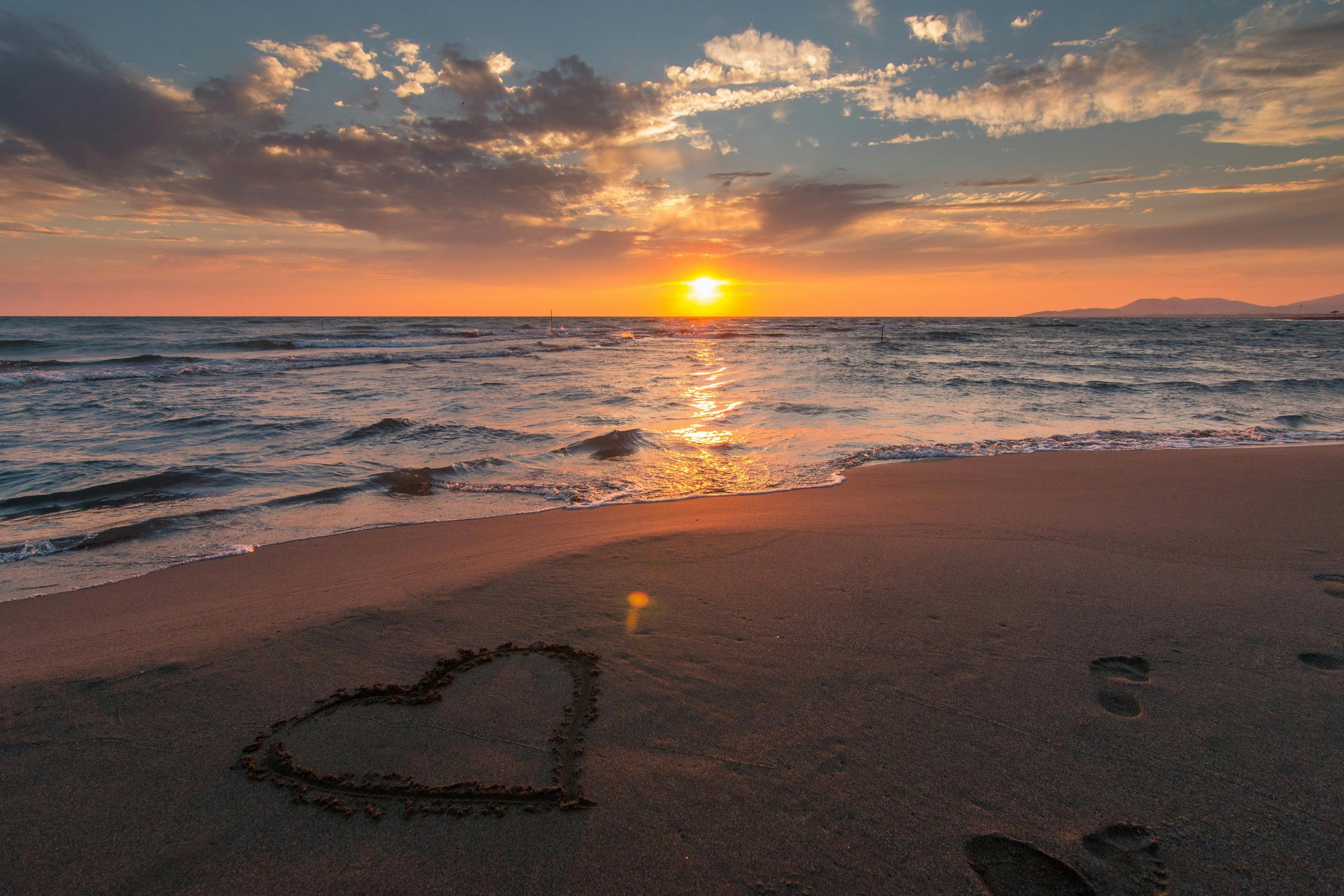 Sunset over the ocean with a sandy beach in the foreground. A heart shape is drawn in the sand, and footprints lead towards the water.