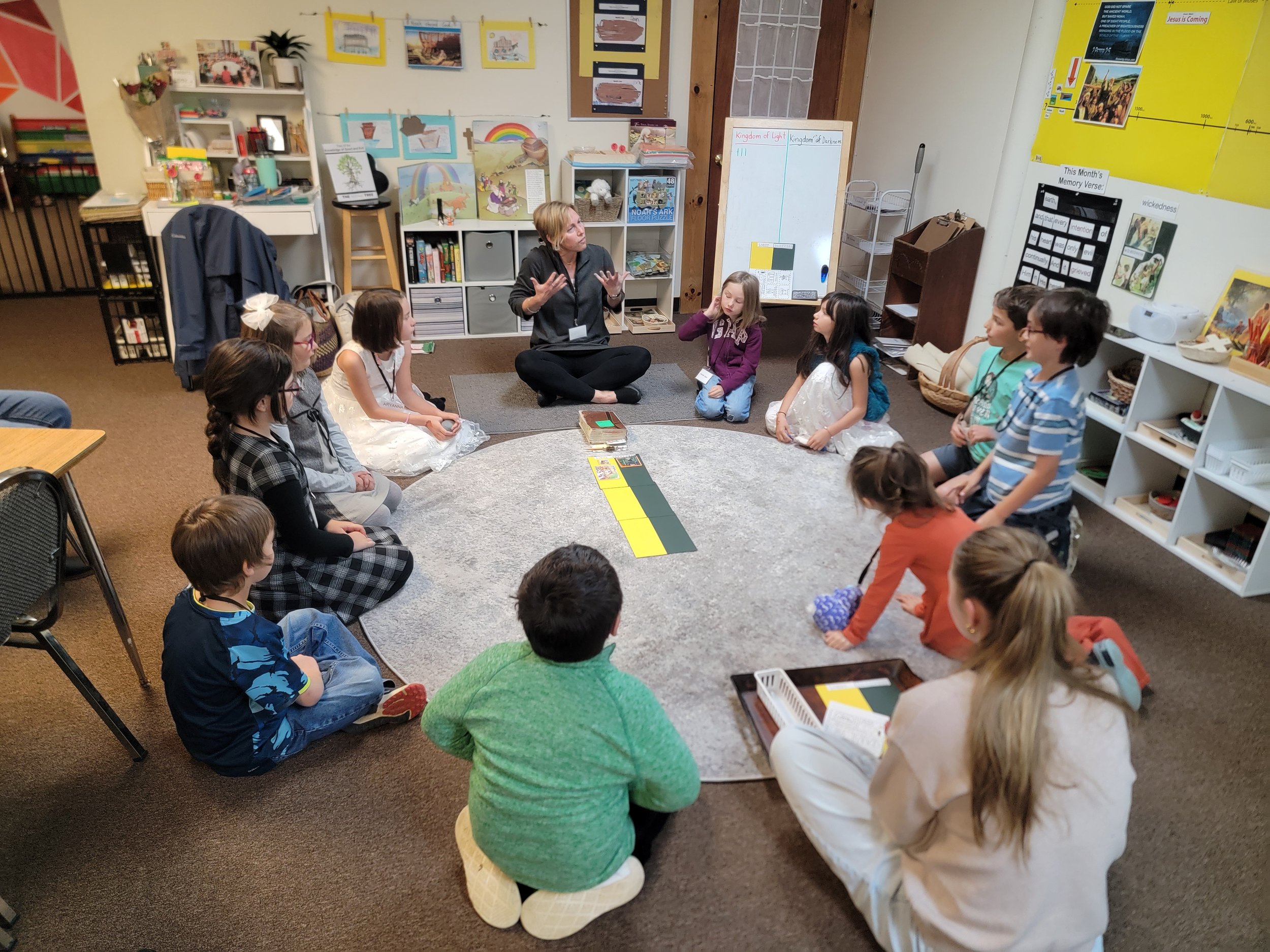A group of children sitting in a circle on a rug around a woman in a classroom. The woman appears to be teaching or leading a discussion. The classroom has books, educational posters, and artwork on shelves and walls.