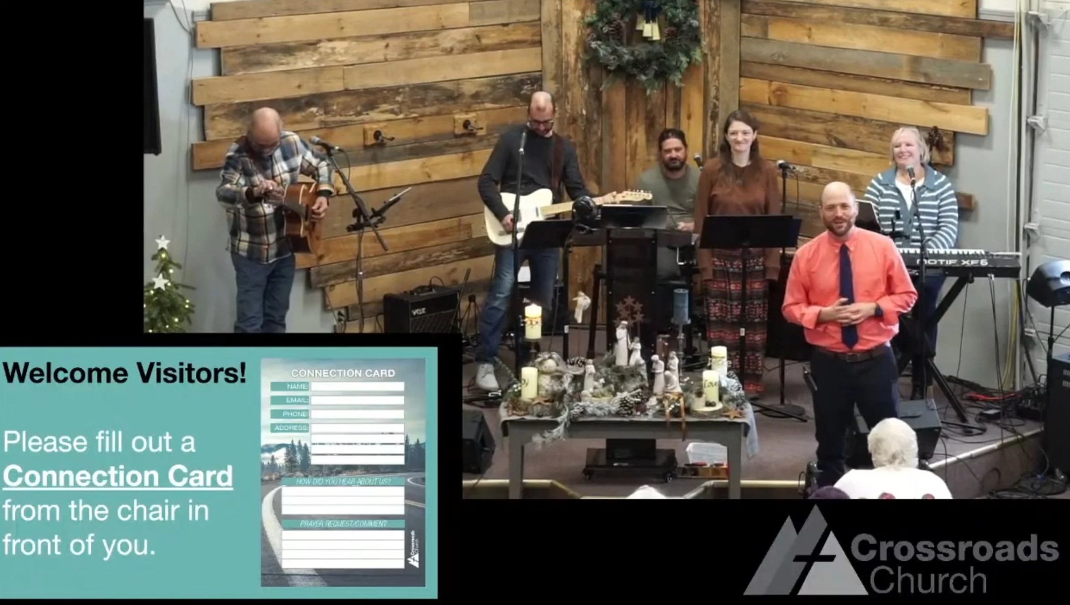 A church service in progress with six musicians on stage, including a person in an orange shirt with a blue tie standing in front of the stage. Decorations include a small Christmas tree, candles, and nativity figures. A large screen displays a welcome message and a connection card form. The church's logo 'Crossroads Church' is in the bottom right corner.