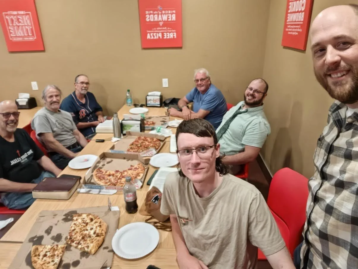 Group of seven men sitting around a table with pizza boxes, pizza slices, plates, and drinks at a restaurant.