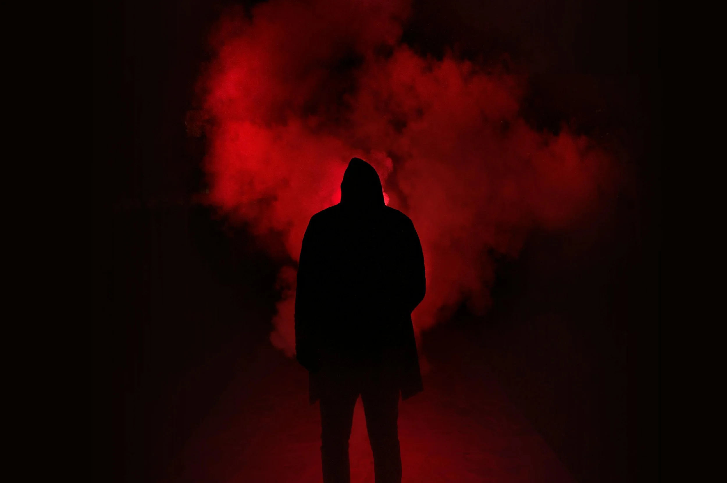 Silhouette of a person in a hoodie standing in front of red smoke in a dark environment.