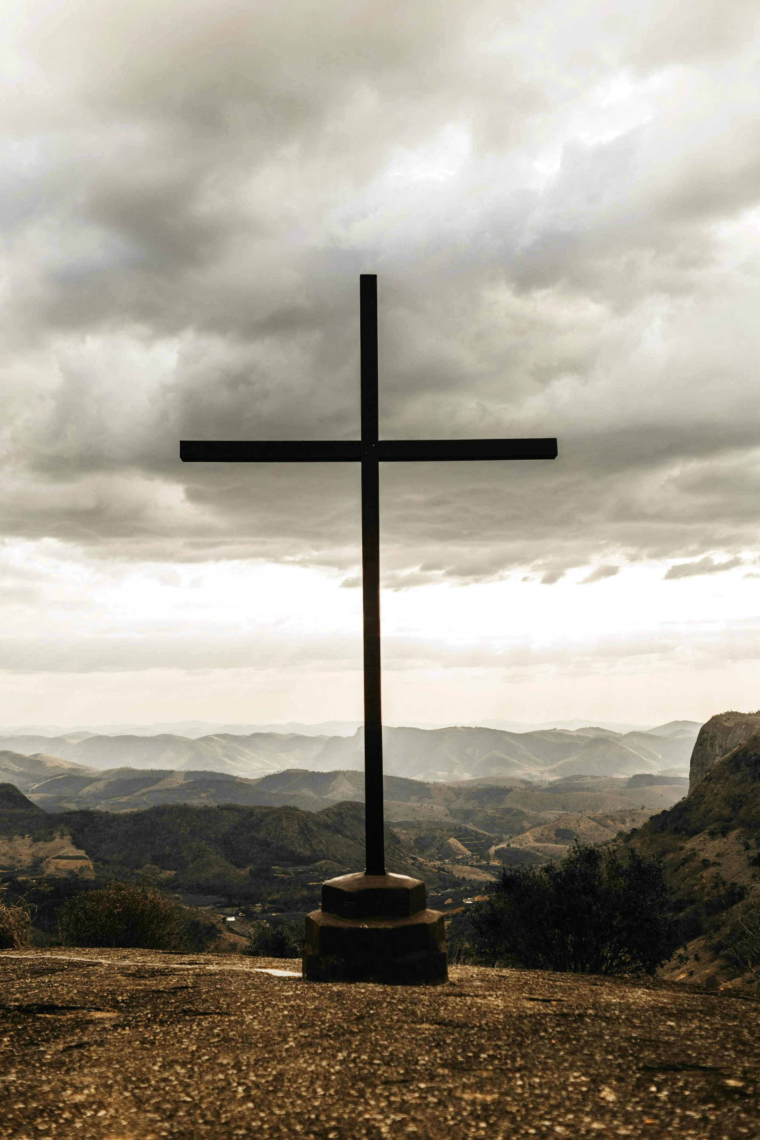 A black cross statue on a concrete base with a mountainous landscape and cloudy sky in the background.
