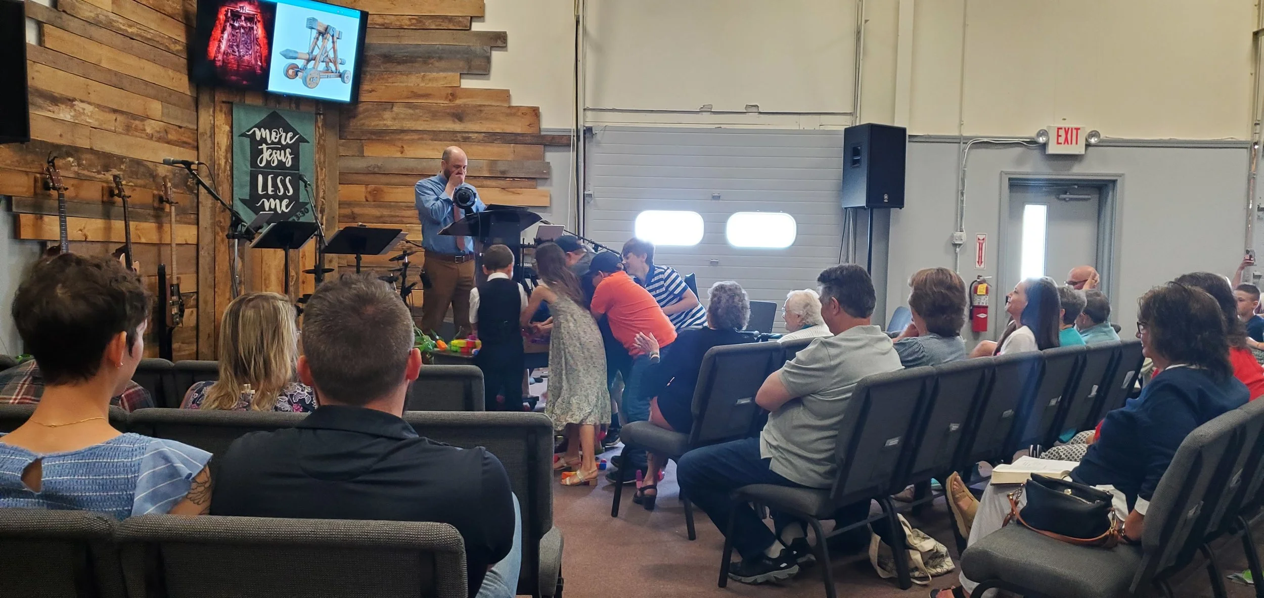 A man is taking a photograph in front of an audience seated in chairs in a church or event space with wooden wall decor and musical instruments, including guitars and a drum set, visible on the stage.