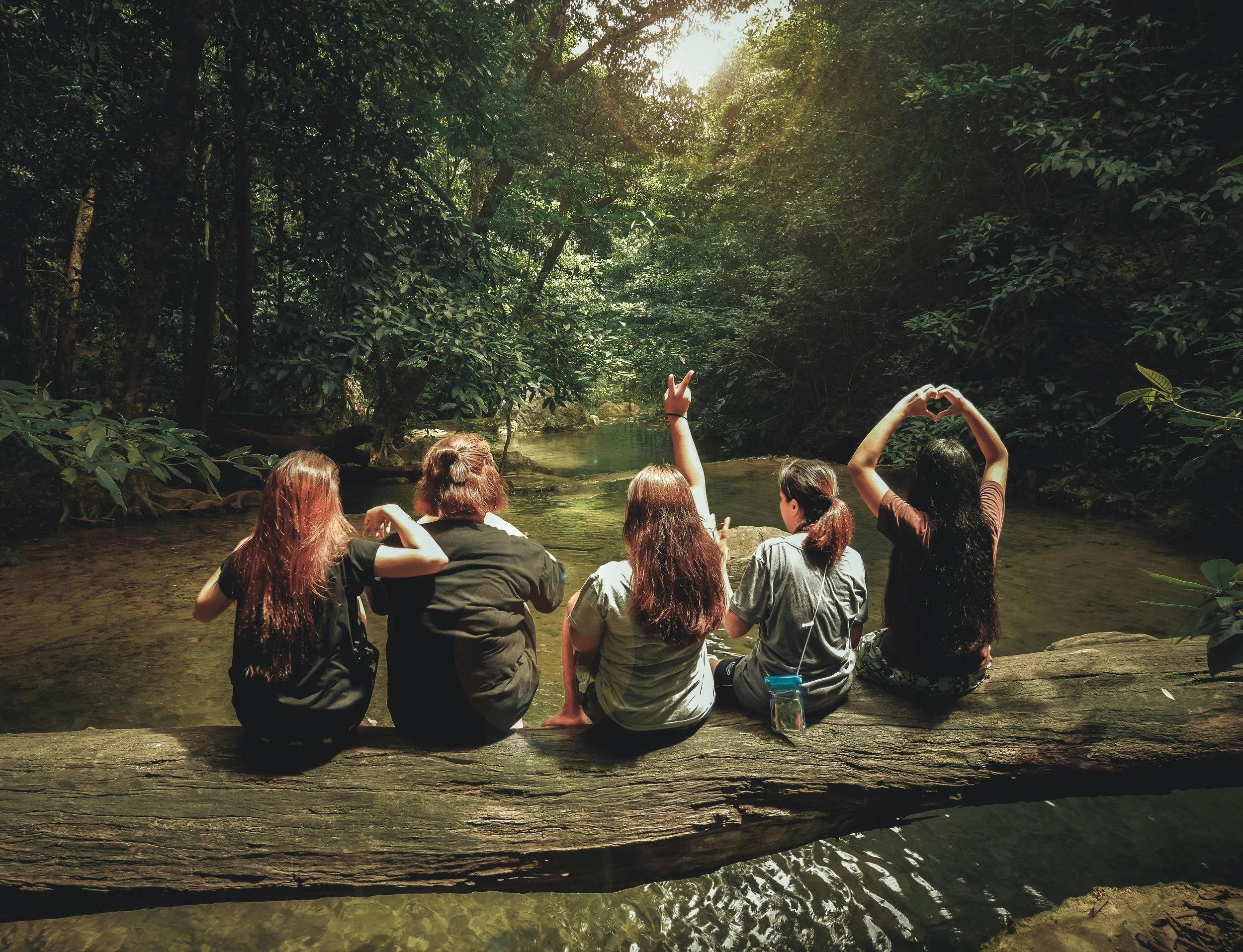 Five girls sitting on a log in a lush forest by a creek, with sunlight filtering through the trees, one making a peace sign and another forming a heart shape with their hands.