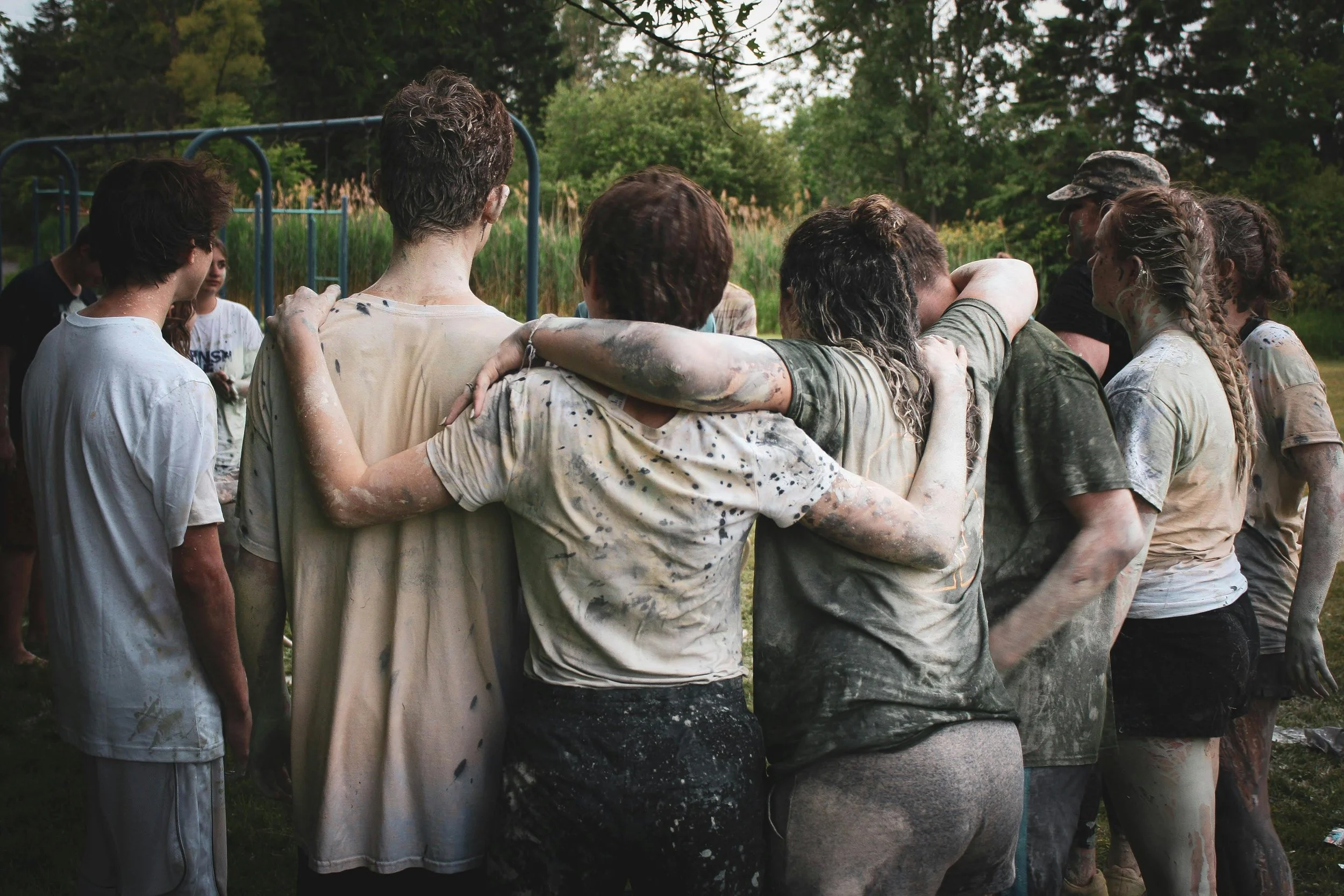 Group of people standing together outdoors with arms around each other, covered in mud, in a park setting.