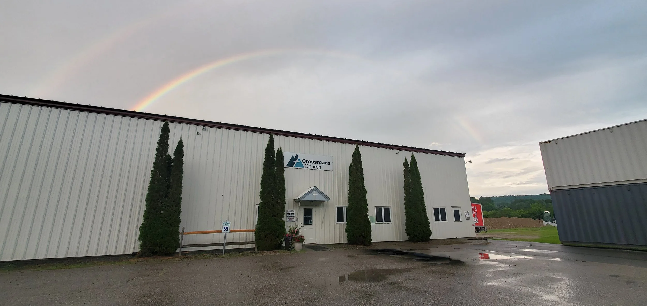 Exterior of Crossroads Church building with four tall bushes in front, and a double rainbow in the cloudy sky above.