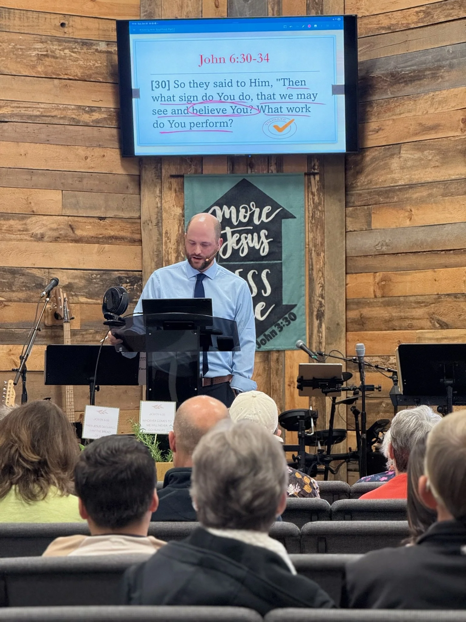 A man leading a church service in a church with wooden walls, standing behind a lectern with a microphone, a large screen displaying a Bible verse from John 6:30-34, and a banner that reads 'more Jesus' on a green background.