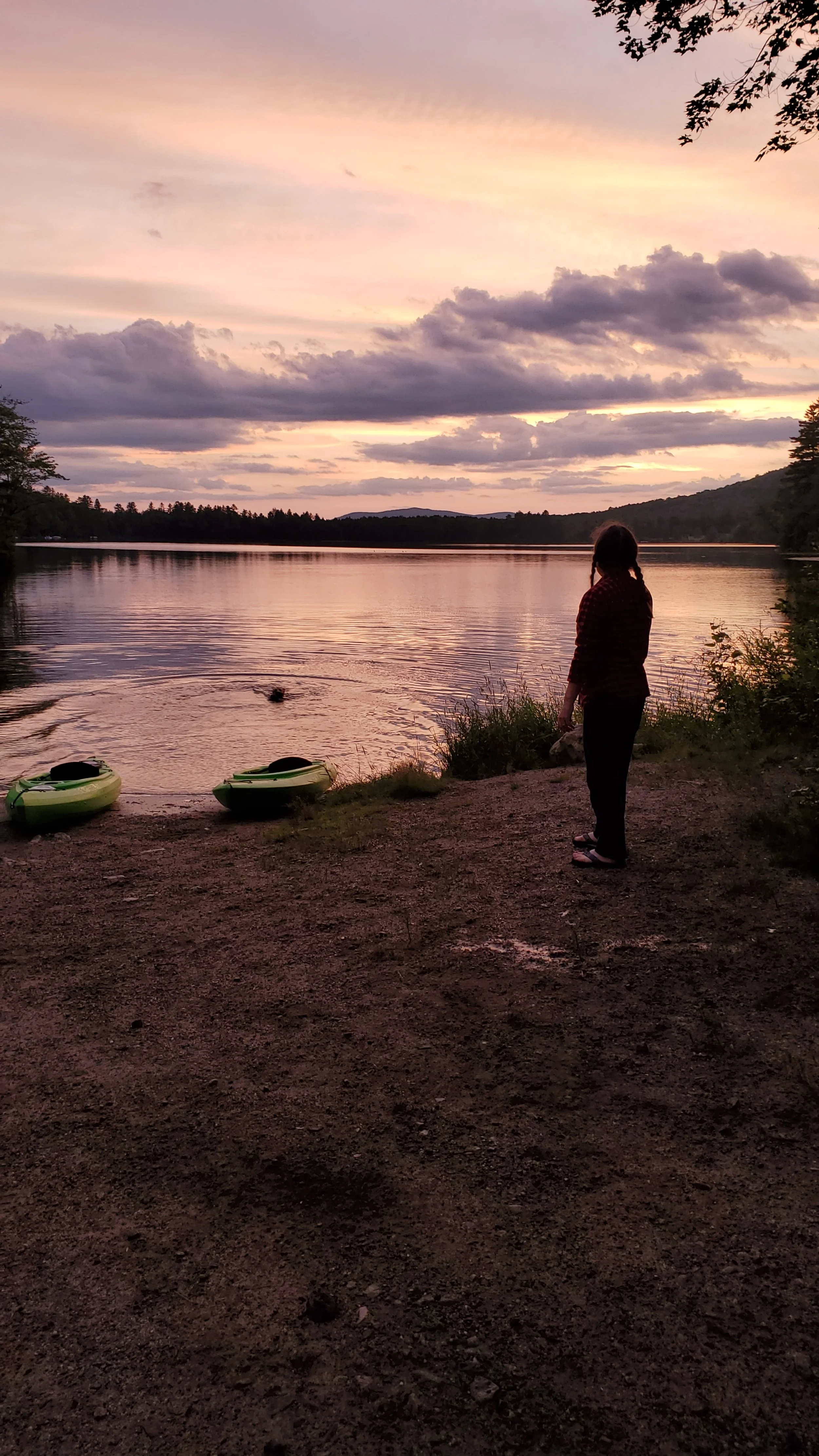 A woman standing on a dirt shore by a lake during sunset, with kayaks on the shore and trees on the sides, cloudy sky reflecting in the water.