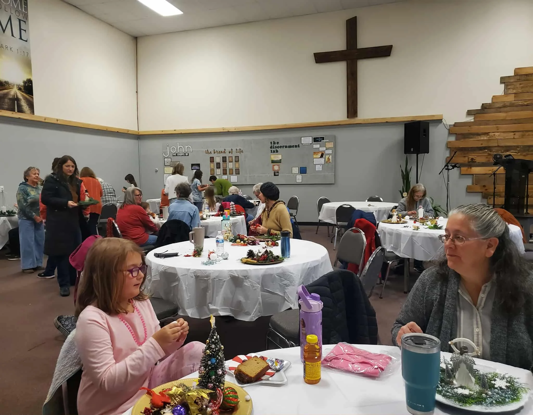 People gathered in a church hall decorated for a Christmas celebration, with tables covered in holiday decorations and food, and a cross on the wall behind.