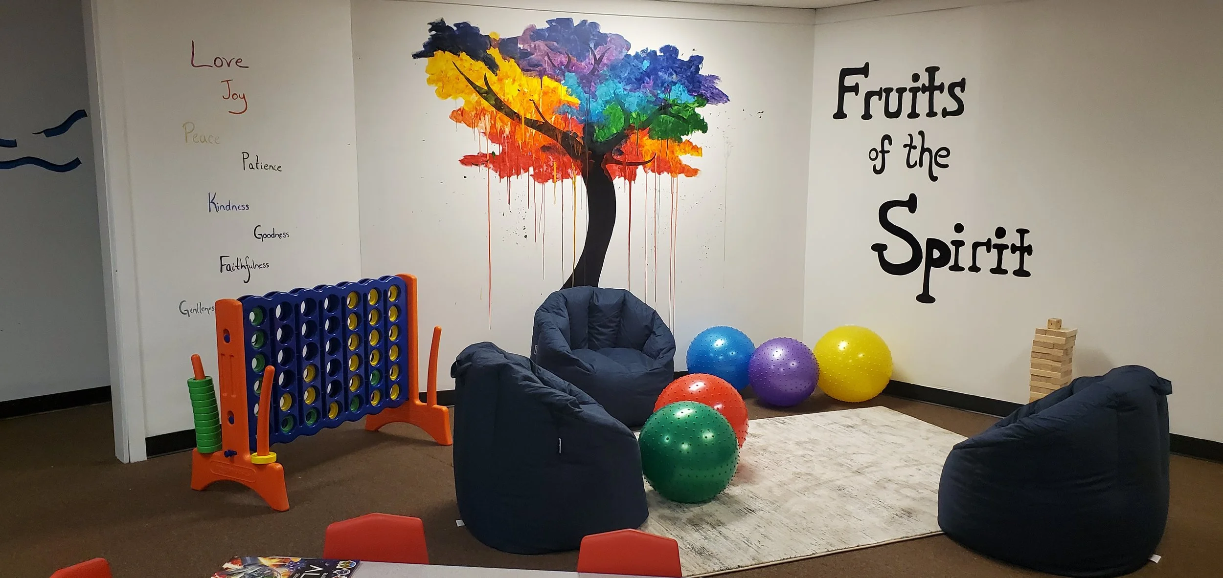 Indoor playroom with a colorful painted tree on the wall, exercise balls in various colors, bean bag chairs, a rectangular rug, and wall art with suns and a list of positive qualities.