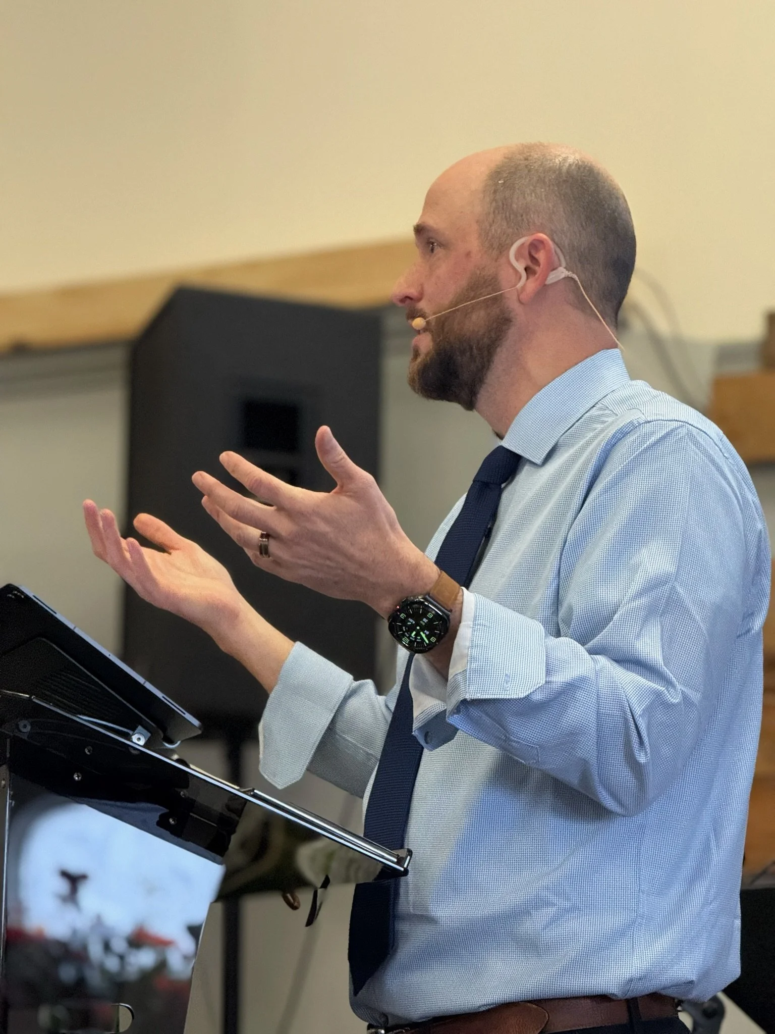 Man with a beard and glasses giving a presentation, wearing a blue shirt, dark tie, and a smartwatch, with a microphone headset and a tablet on a stand.