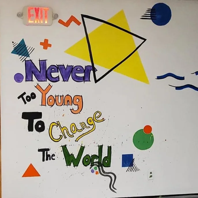 Colorful hand-drawn motivational quote on a white wall, reading 'Never Too Young To Change The World,' decorated with various geometric shapes and patterns.