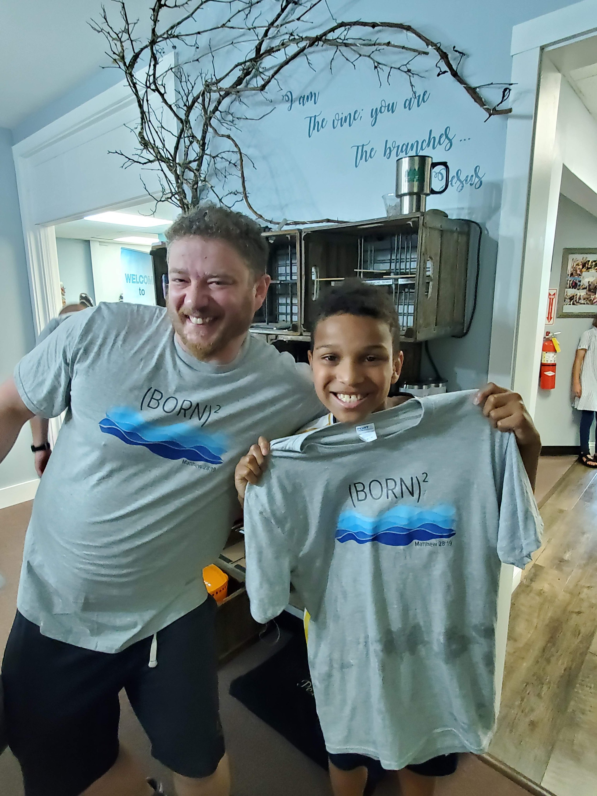 A man and boy smiling and holding matching T-shirts with a wave design and the phrase '(BORN)^2' with Matthew 28:19 written on them, standing inside a room with a mural of branches and a quote on the wall behind them.