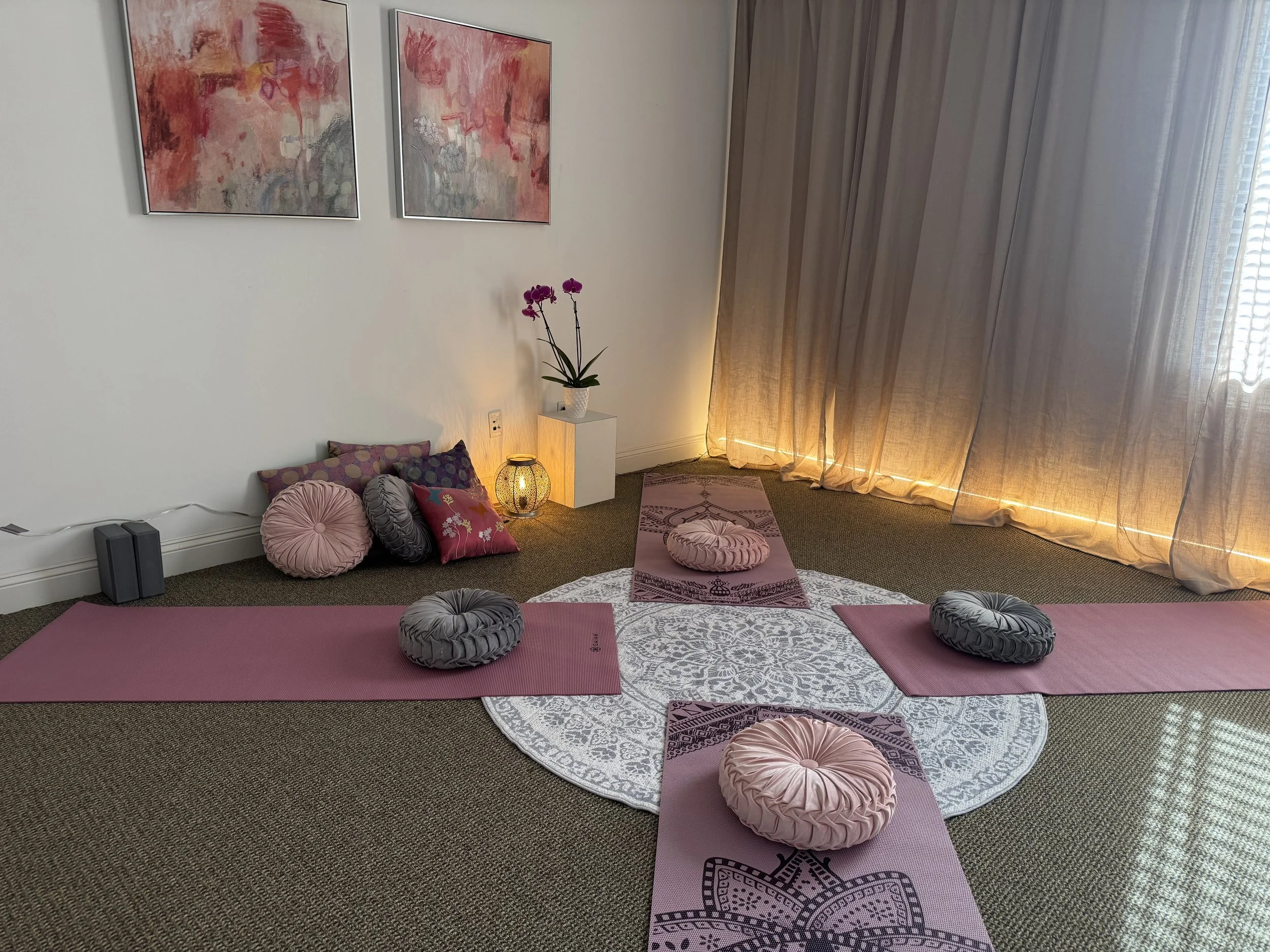 A cozy yoga or meditation space featuring two pink yoga mats with round plush cushions on a patterned circular rug, soft lighting, wall art, pillows, a side table with an orchid plant, and sheer curtains.
