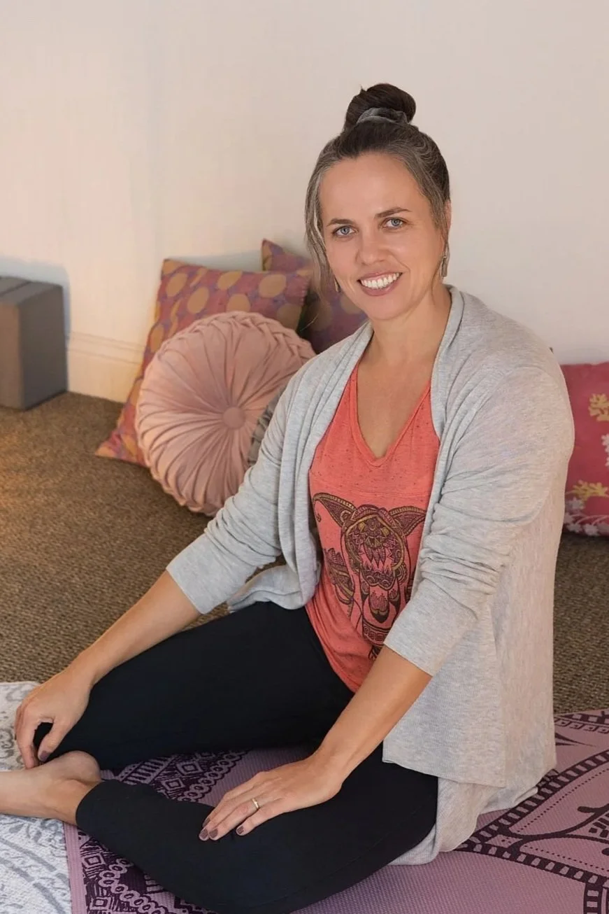 A woman with gray hair tied in a bun, smiling and sitting cross-legged on a yoga mat indoors, in front of colorful pillows on a bed. She is wearing a red T-shirt with a tiger design and a light gray cardigan.