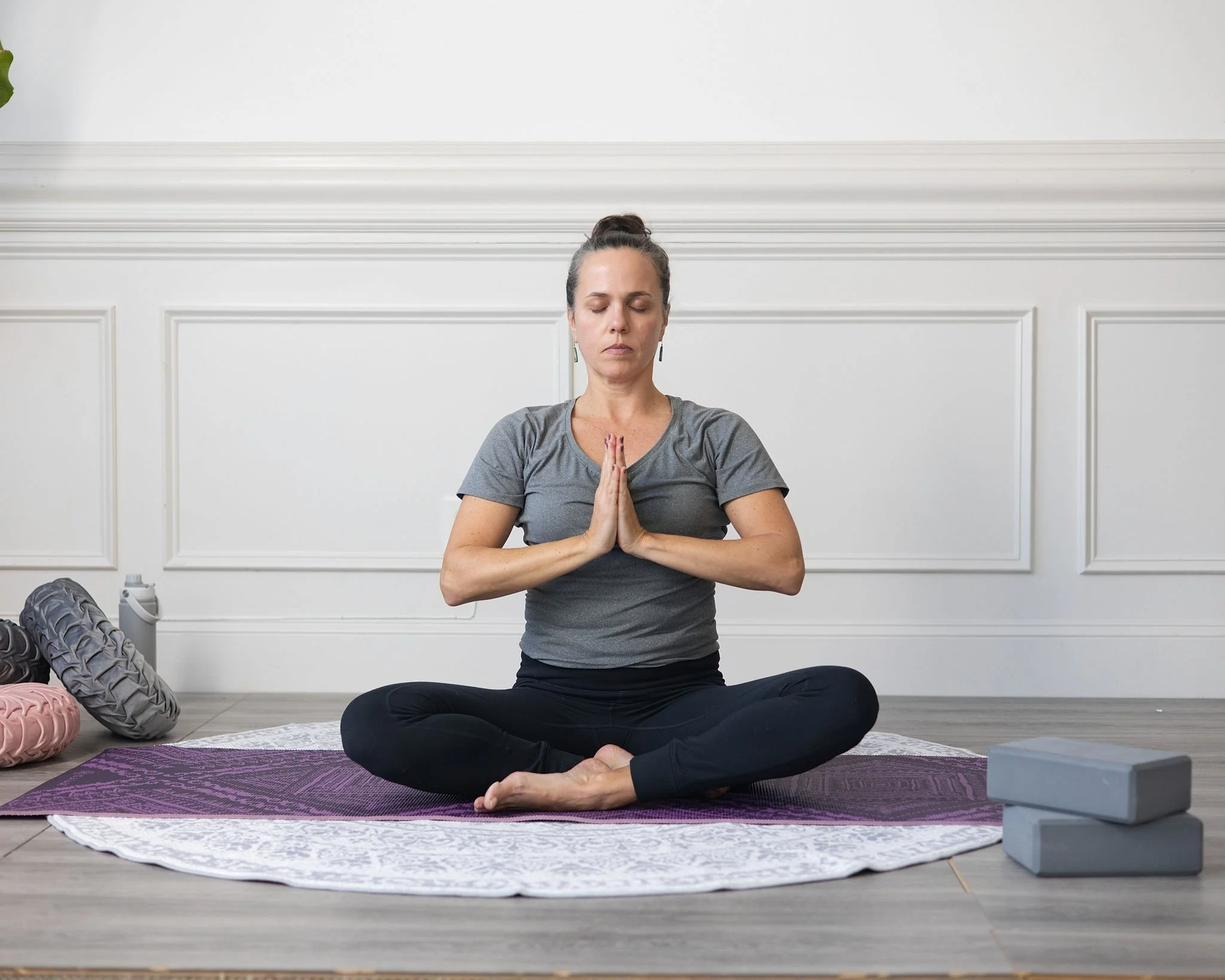 Woman practicing yoga in a cross-legged seated position on a purple yoga mat, with her hands in prayer position, in a room with white walls and yoga props nearby.