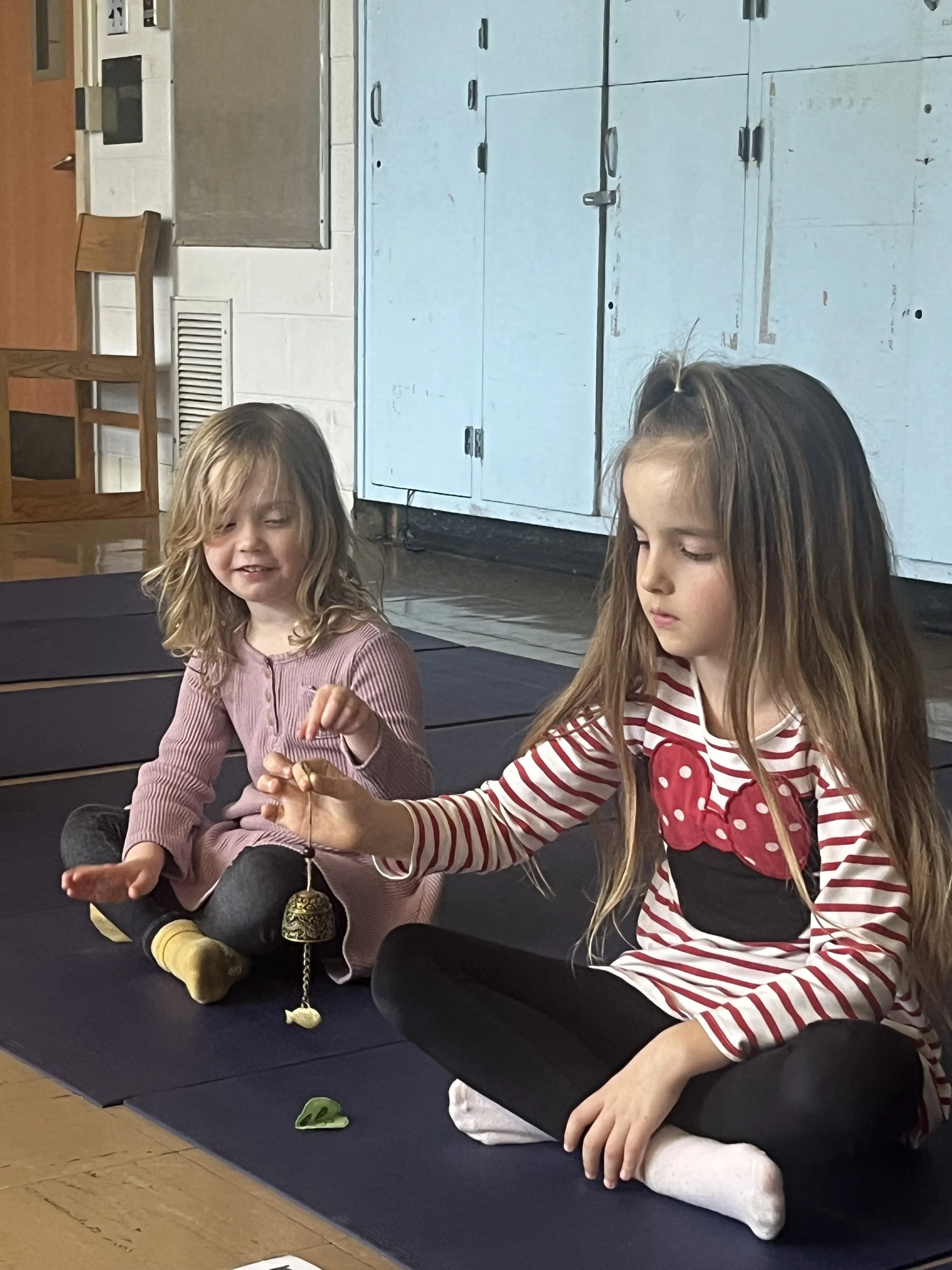 Two young girls sitting on a black mat in a room with blue cabinets, one girl holding a small bell, the other girl watching