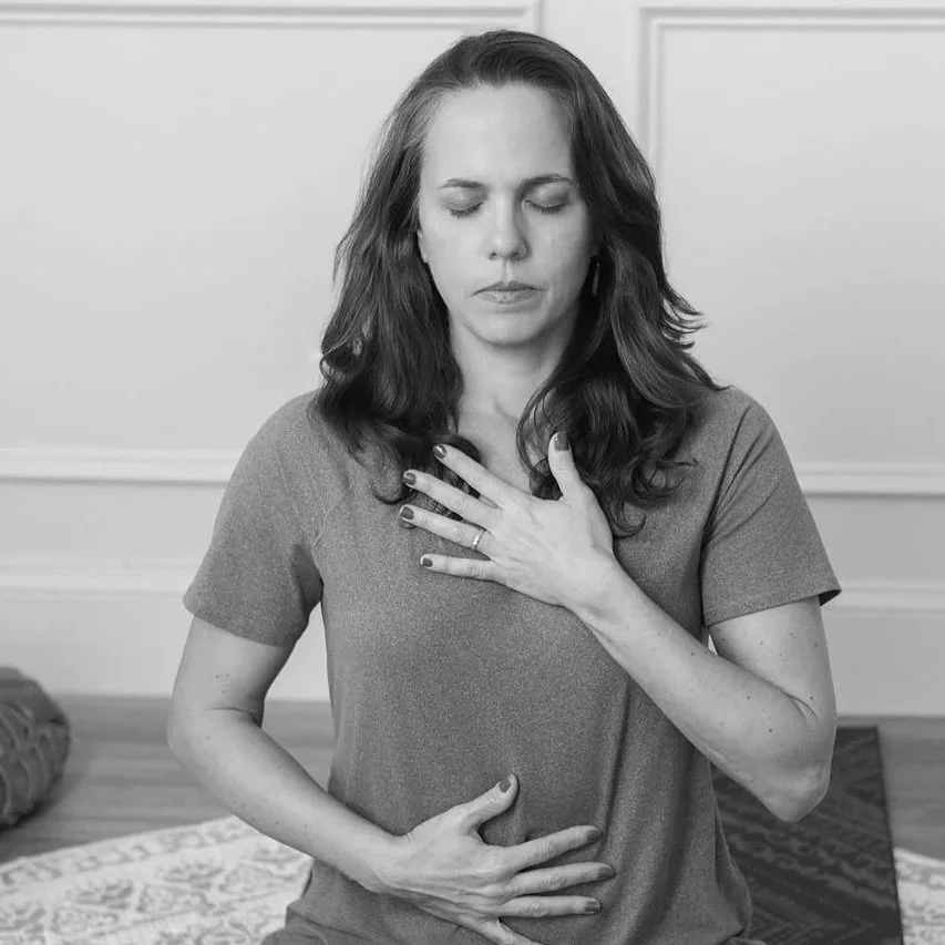 A woman with long wavy hair practicing yoga or meditation, sitting in a cross-legged position, with one hand on her chest and one on her abdomen, eyes closed, in a peaceful indoor setting.