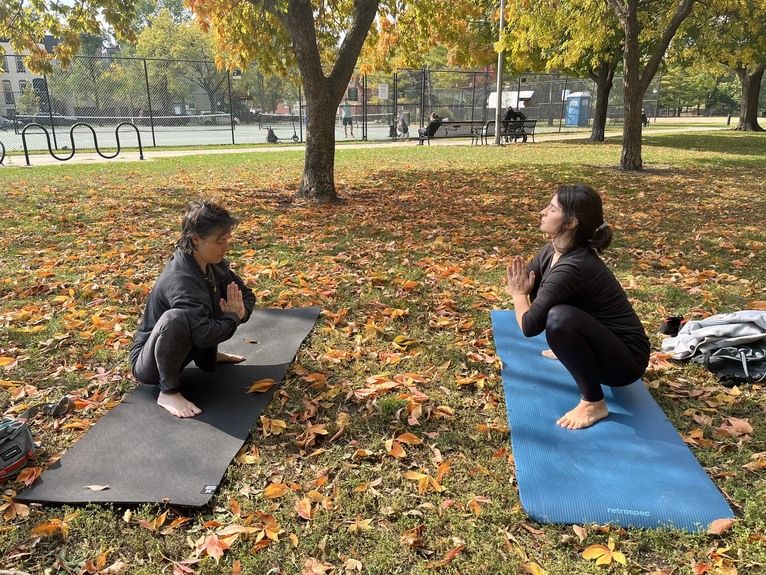 Two women practicing yoga outdoors in a park during fall, kneeling on yoga mats with palms pressed together in a prayer position, surrounded by fallen autumn leaves and trees.