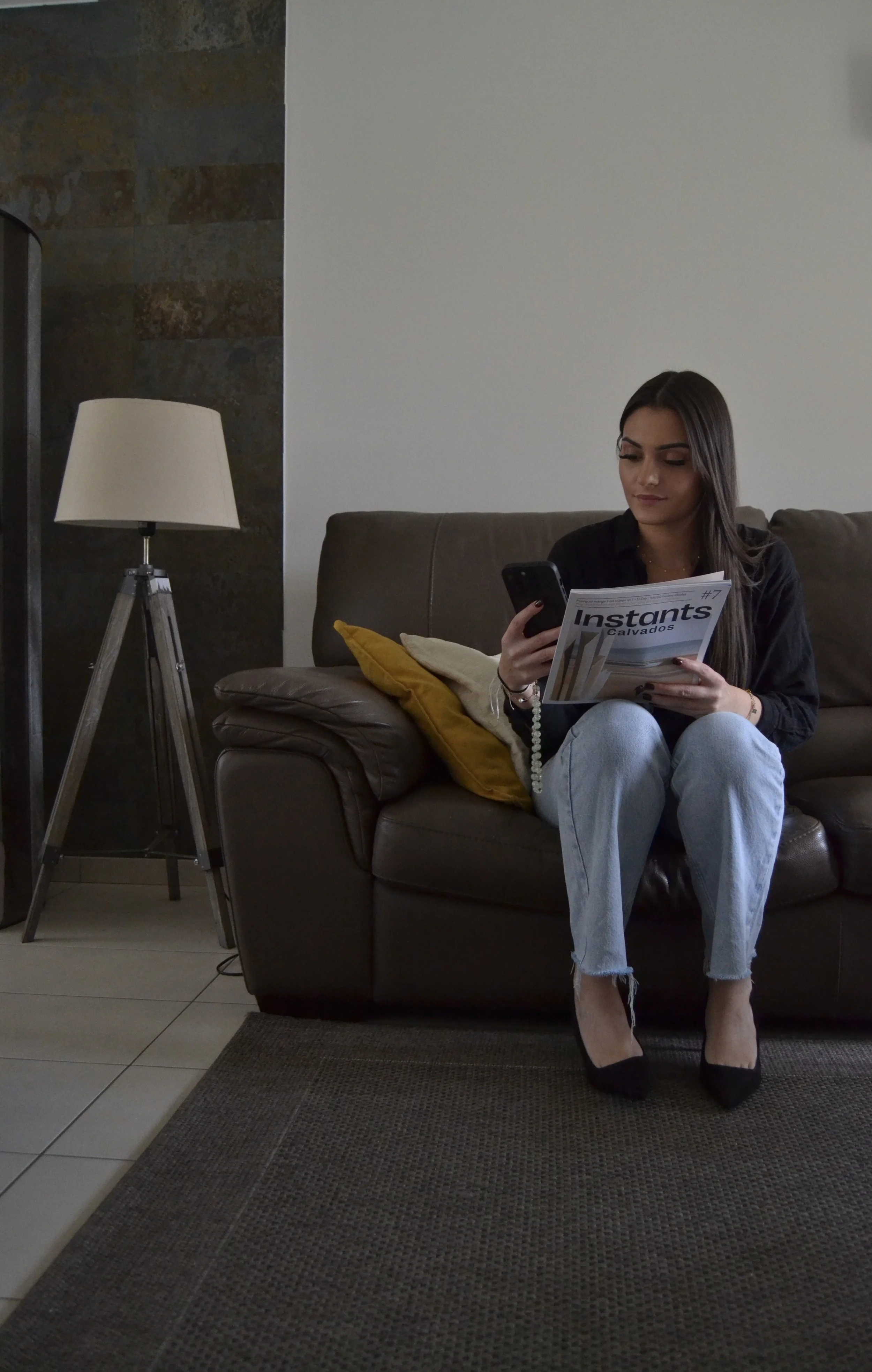 Une femme assise sur un canapé en cuir foncé, tenant un téléphone portable dans une main et un magazine dans l'autre, dans un salon moderne avec une lampe sur pied et un mur blanc.