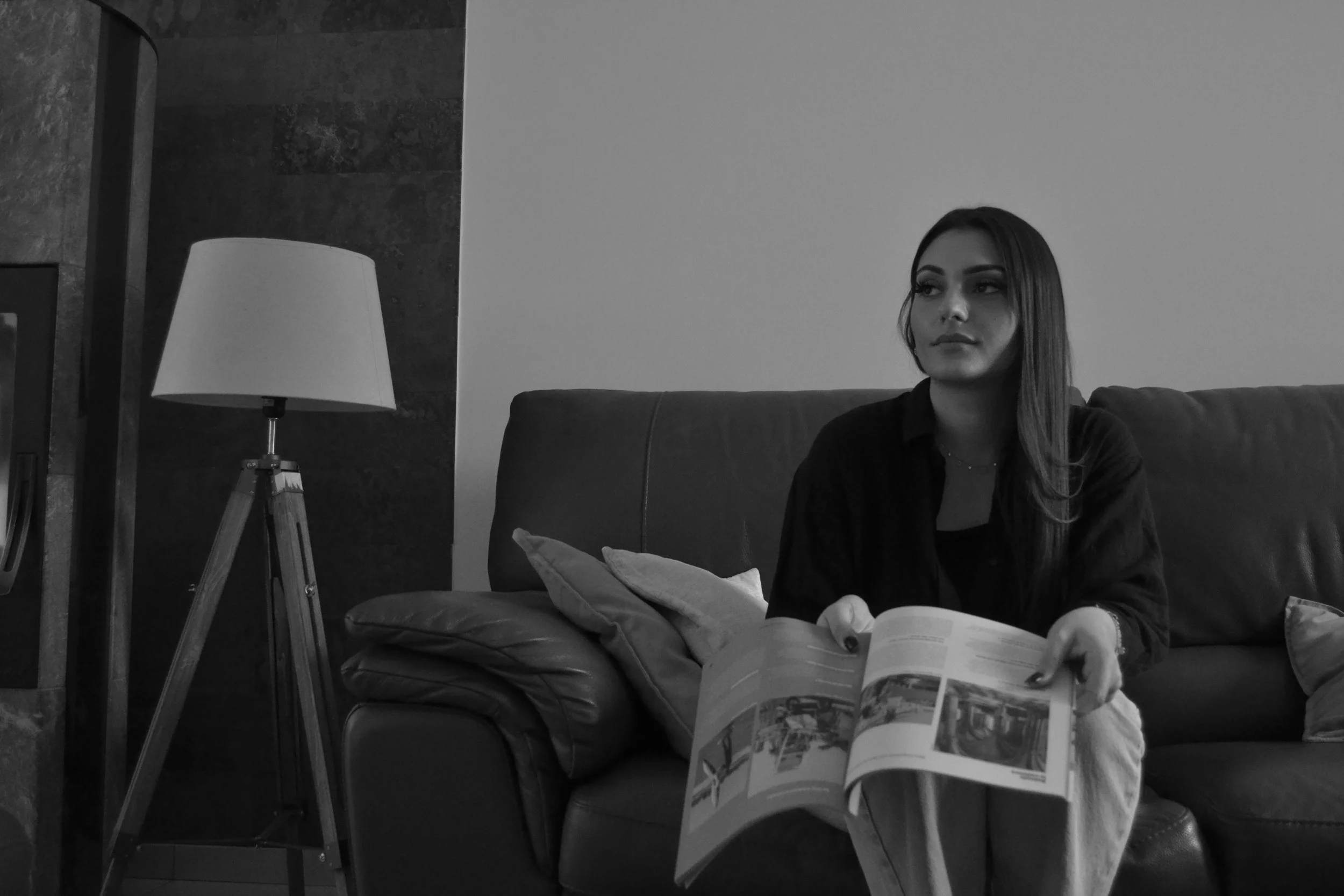 Une femme assise sur un canapé en cuir lit un magazine dans un salon avec une lampe sur un trépied à gauche.