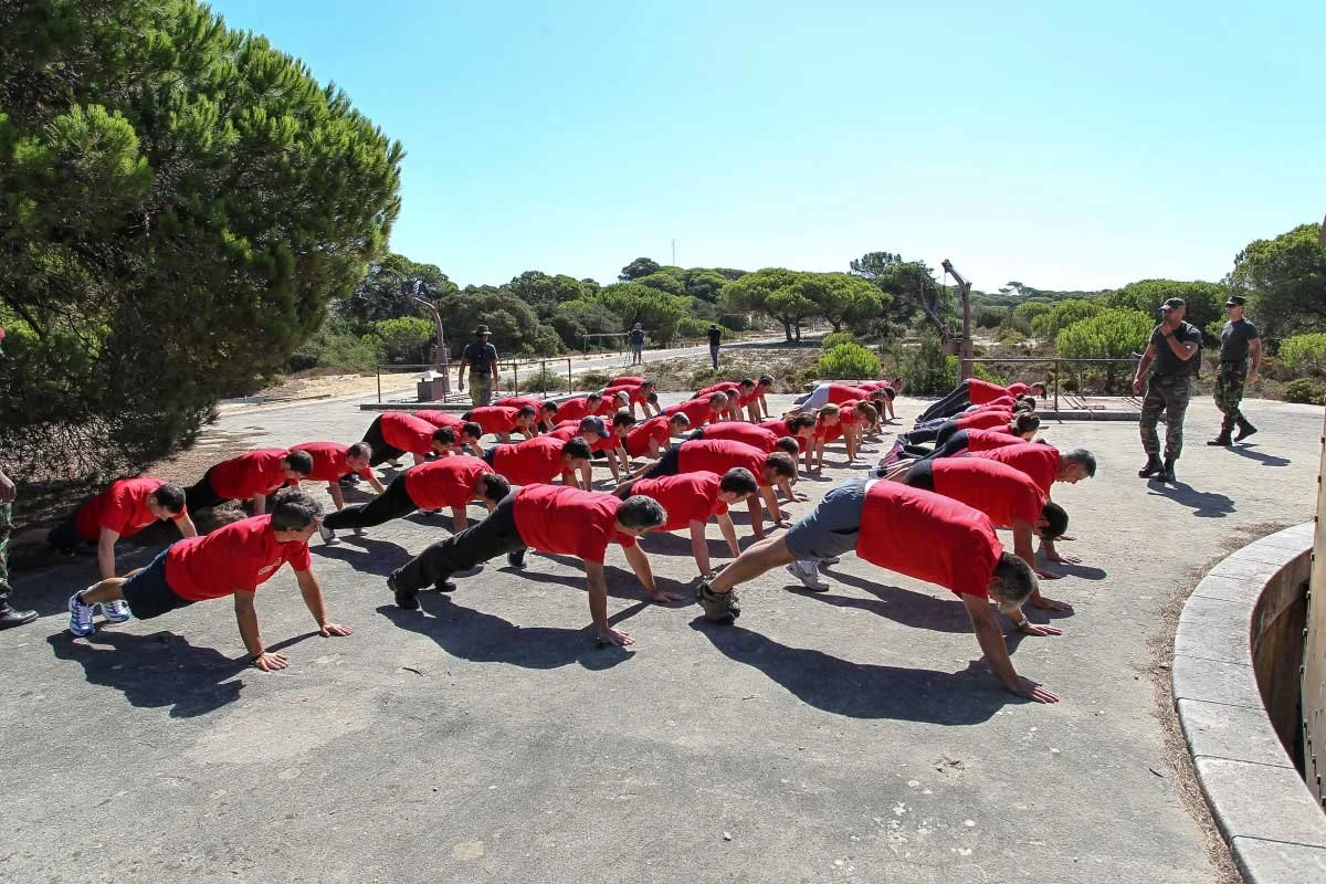 Group of young people doing push-ups outdoors on a sunny day, with some instructors nearby, in a natural setting with trees and bushes.