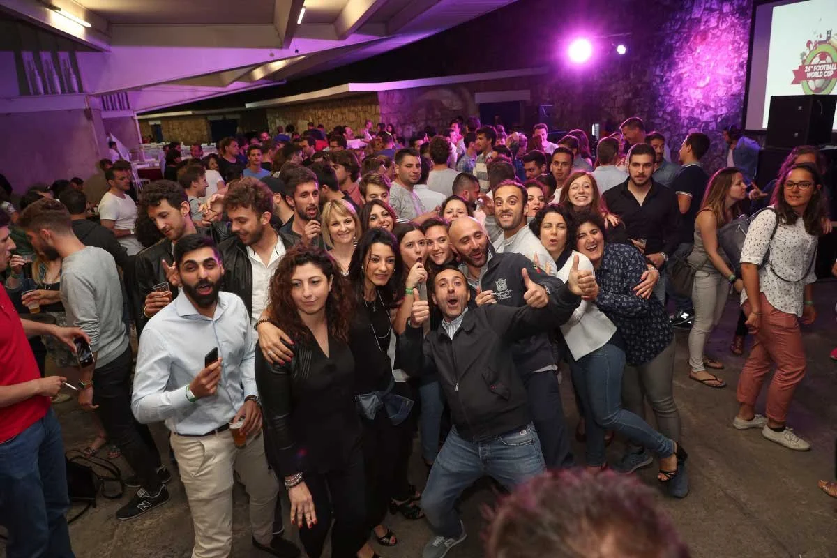 Large group of people at a lively indoor event, some holding drinks, smiling, and posing for a photo with purple lighting and a large screen displaying a logo in the background.