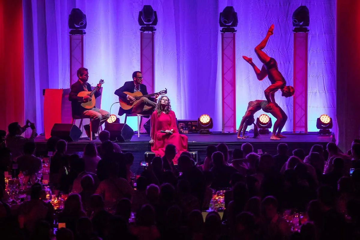 Musicians and dancers on a stage with purple lighting, performing before an audience in a dimly lit room.