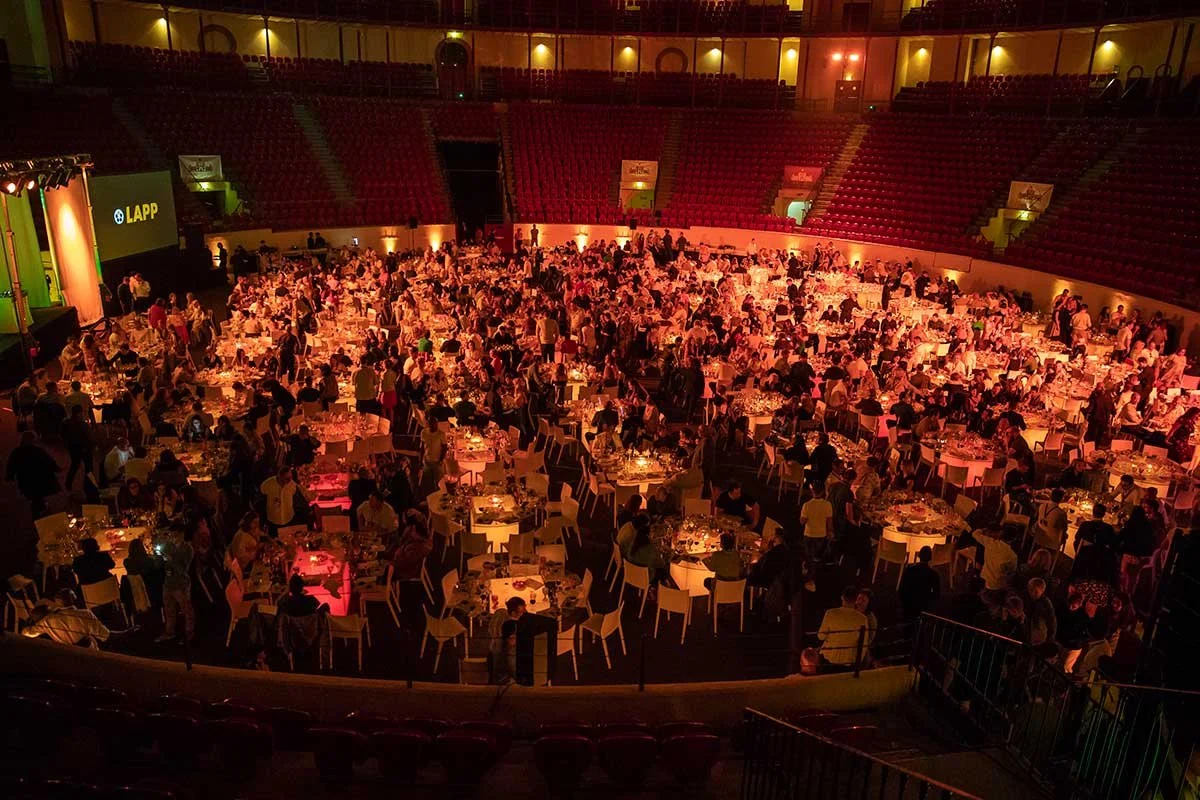A large indoor event with many round tables set for dining, filled with people, in a dimly lit, theater-style venue.