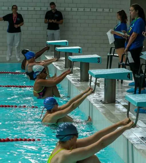Swimmers at a pool starting block during a race, with officials and coaches watching.