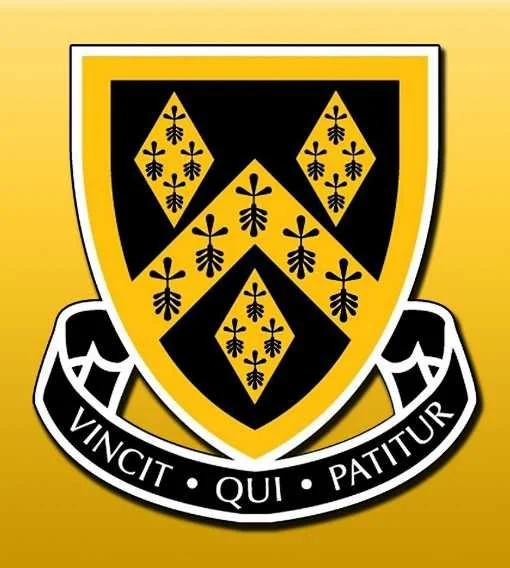 The emblem of a shield with a black and gold color scheme featuring a pattern of gold fleur-de-lis symbols, and a banner below with the Latin phrase 'Vincit Qui Patitur'.