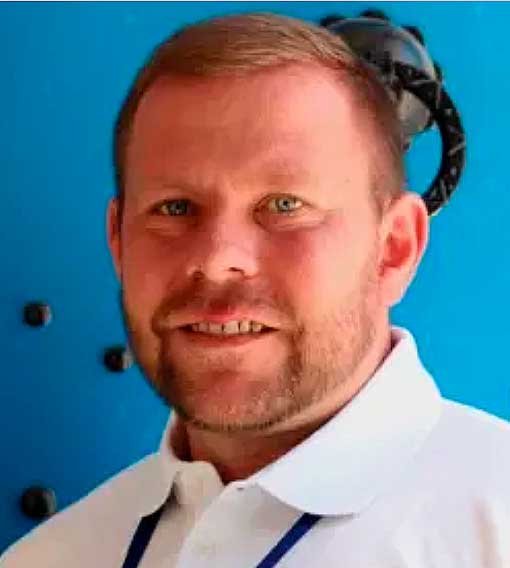 A man with light skin, short reddish-brown hair, and a beard, wearing a white collared shirt, standing in front of a blue background with a black object behind his head.