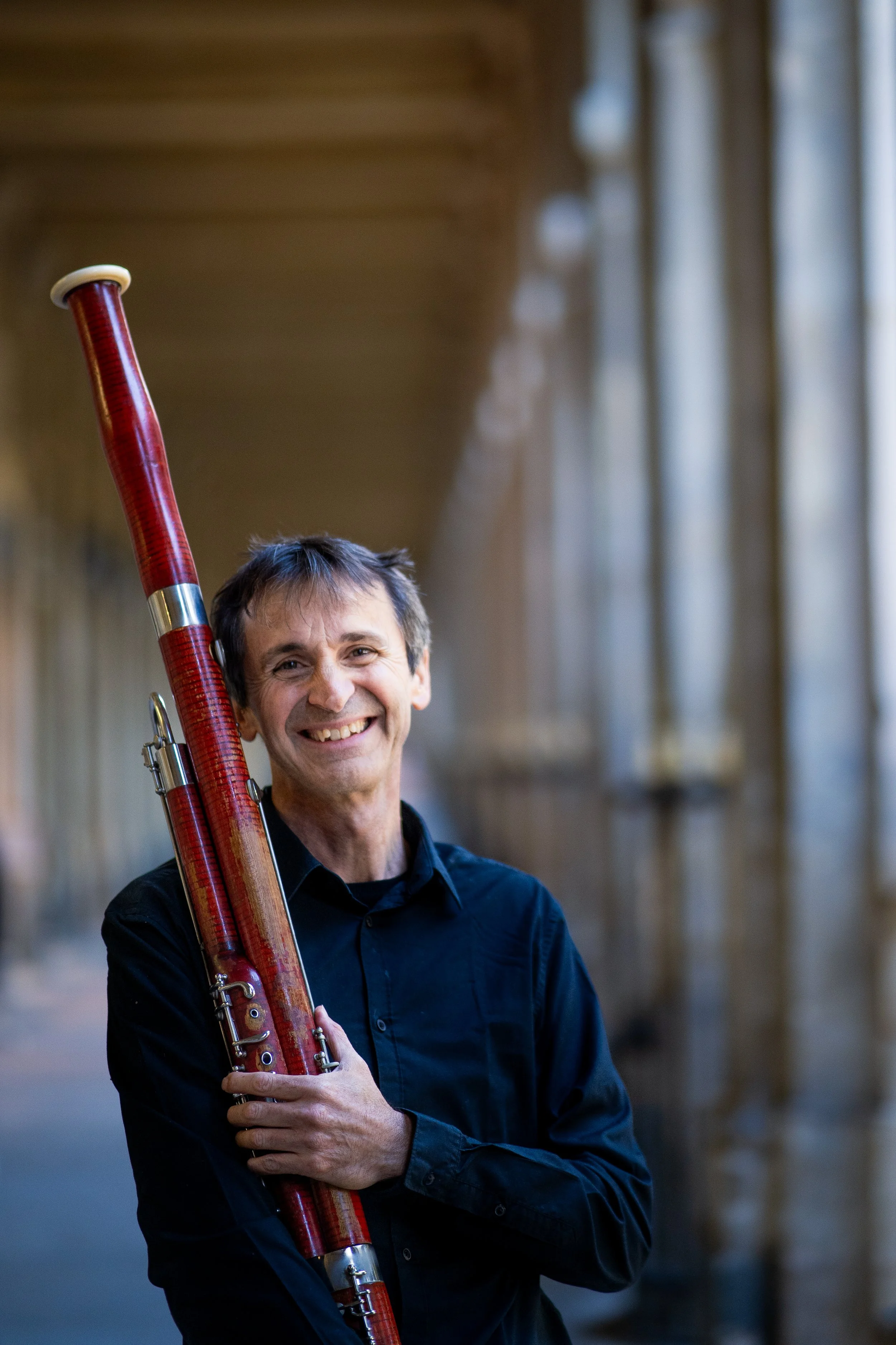 Hombre sosteniendo una fagot en un corredor con columnas de piedra.