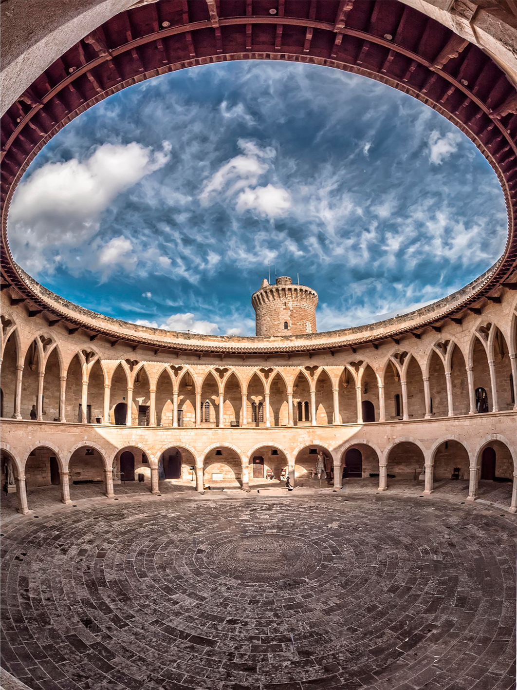Vista del interior de un castillo con patio circular, torres y arco en la estructura, cielo con nubes arriba.
