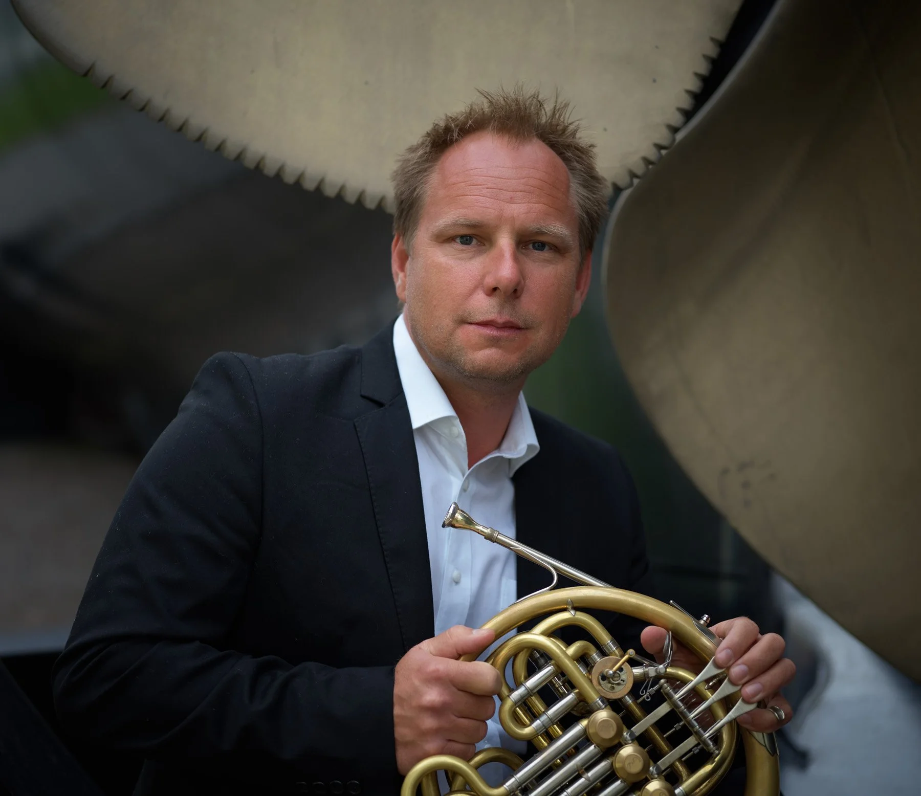 Hombre con cabello despeinado, vestido con saco negro y camisa blanca, sosteniendo un instrumento musical de viento en un entorno al aire libre.