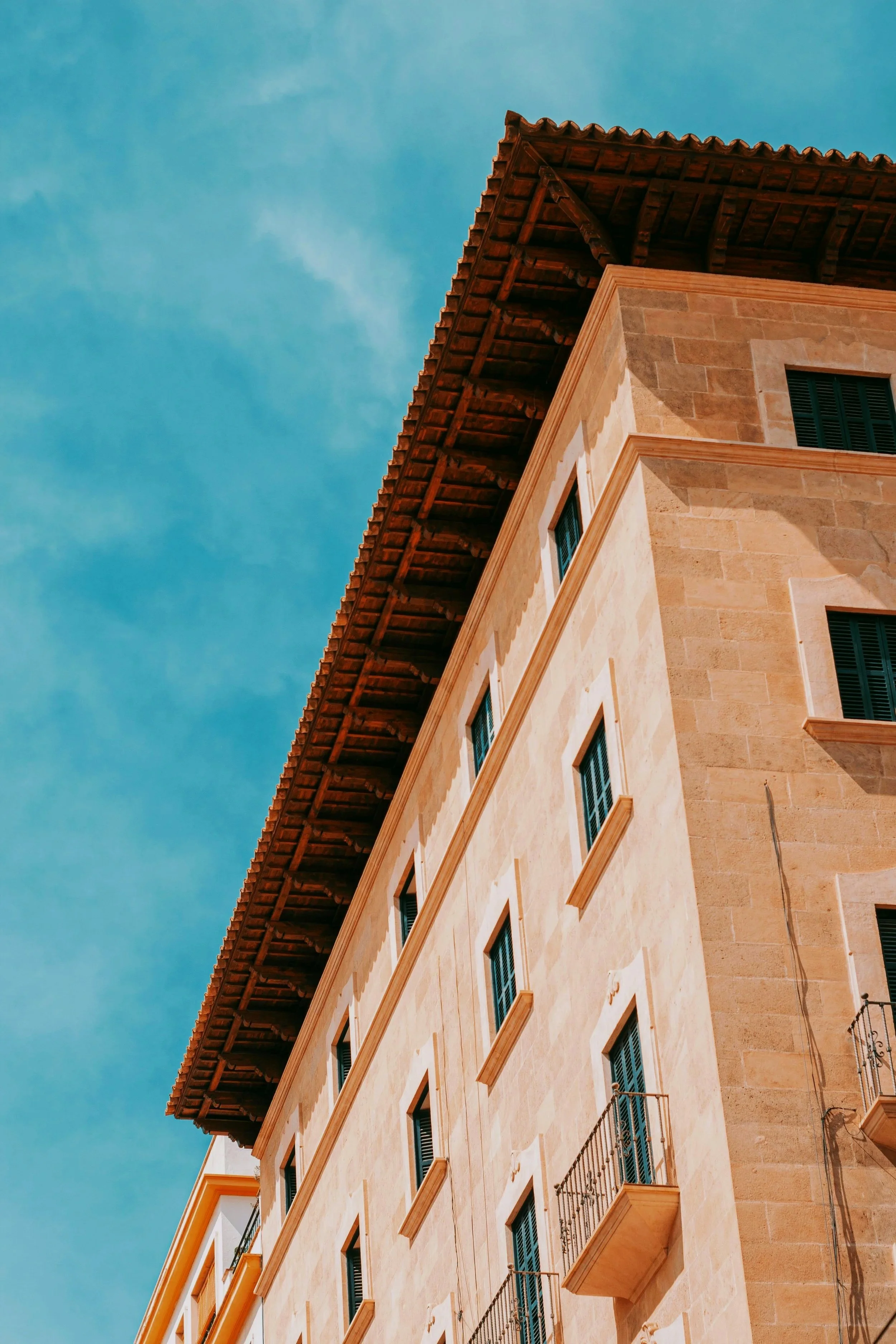 Edificio de piedra con ventanas y balcones pequeños, con un techo de tejas, bajo un cielo azul.