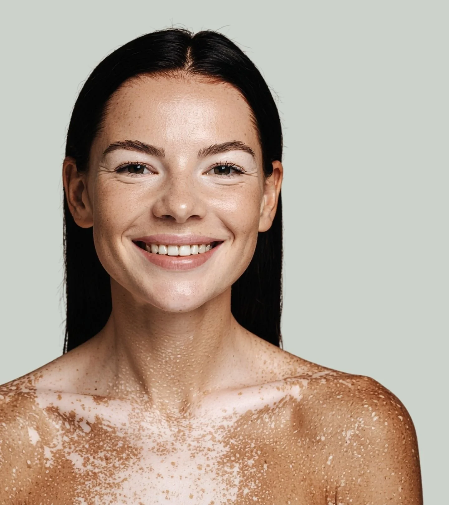 A smiling woman with black hair and freckles on her face and shoulders, with some on her chest, against a plain light gray background.