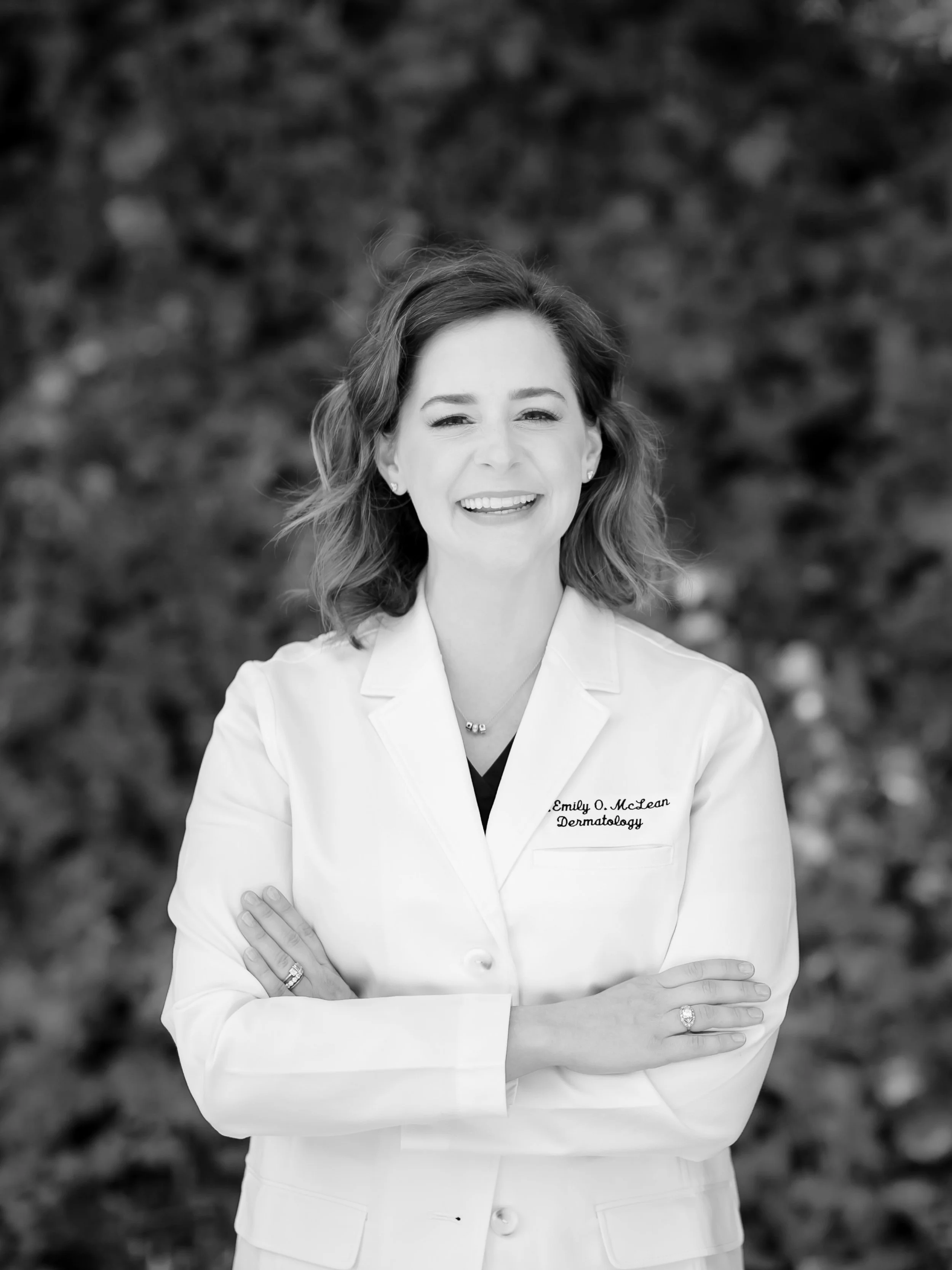 Black and white photo of a woman in a medical coat with her arms crossed, standing outside in front of a blurred natural background.
