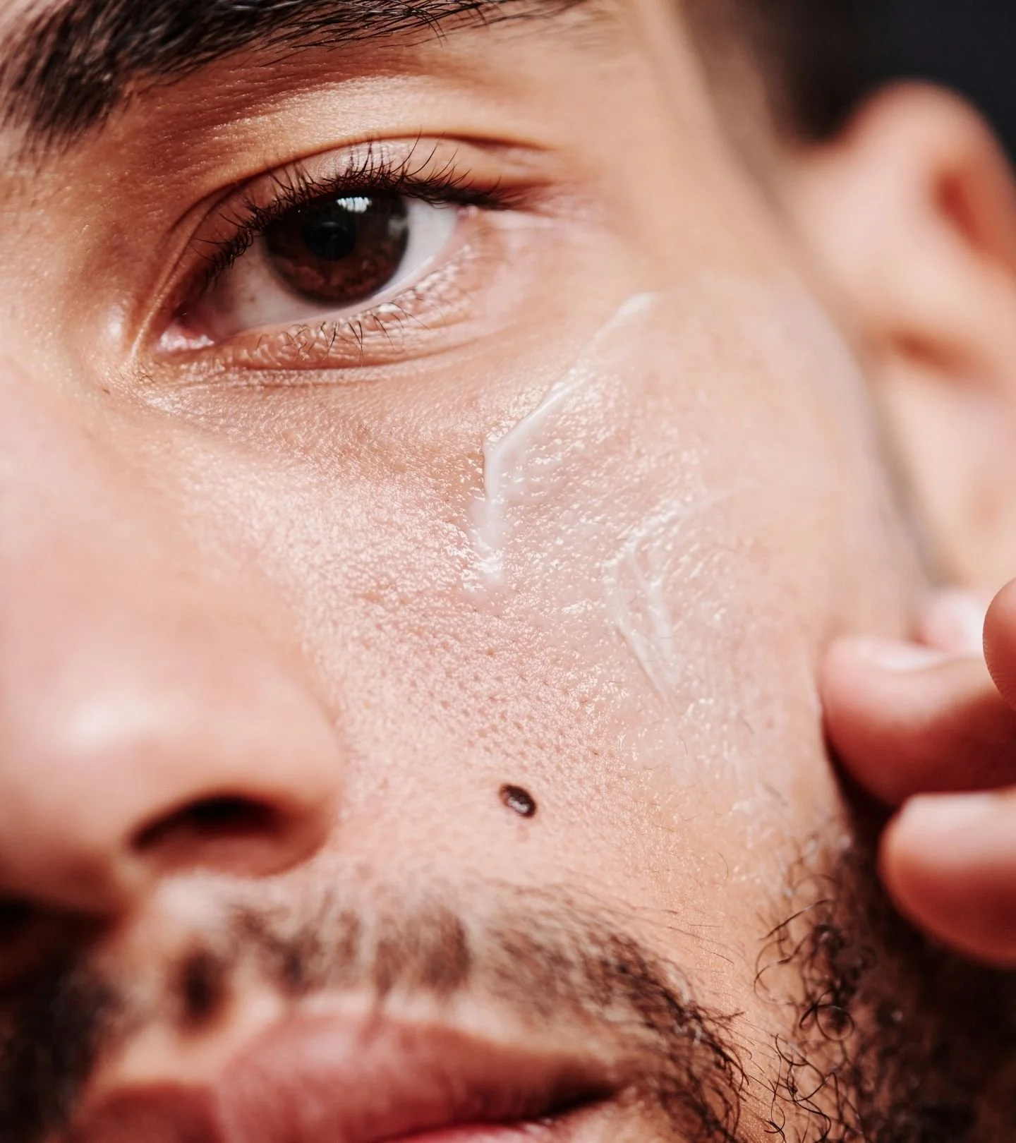 Close-up of a man's face, showing eye, nose, and beard; with a clear strip of skincare product applied on his cheek.