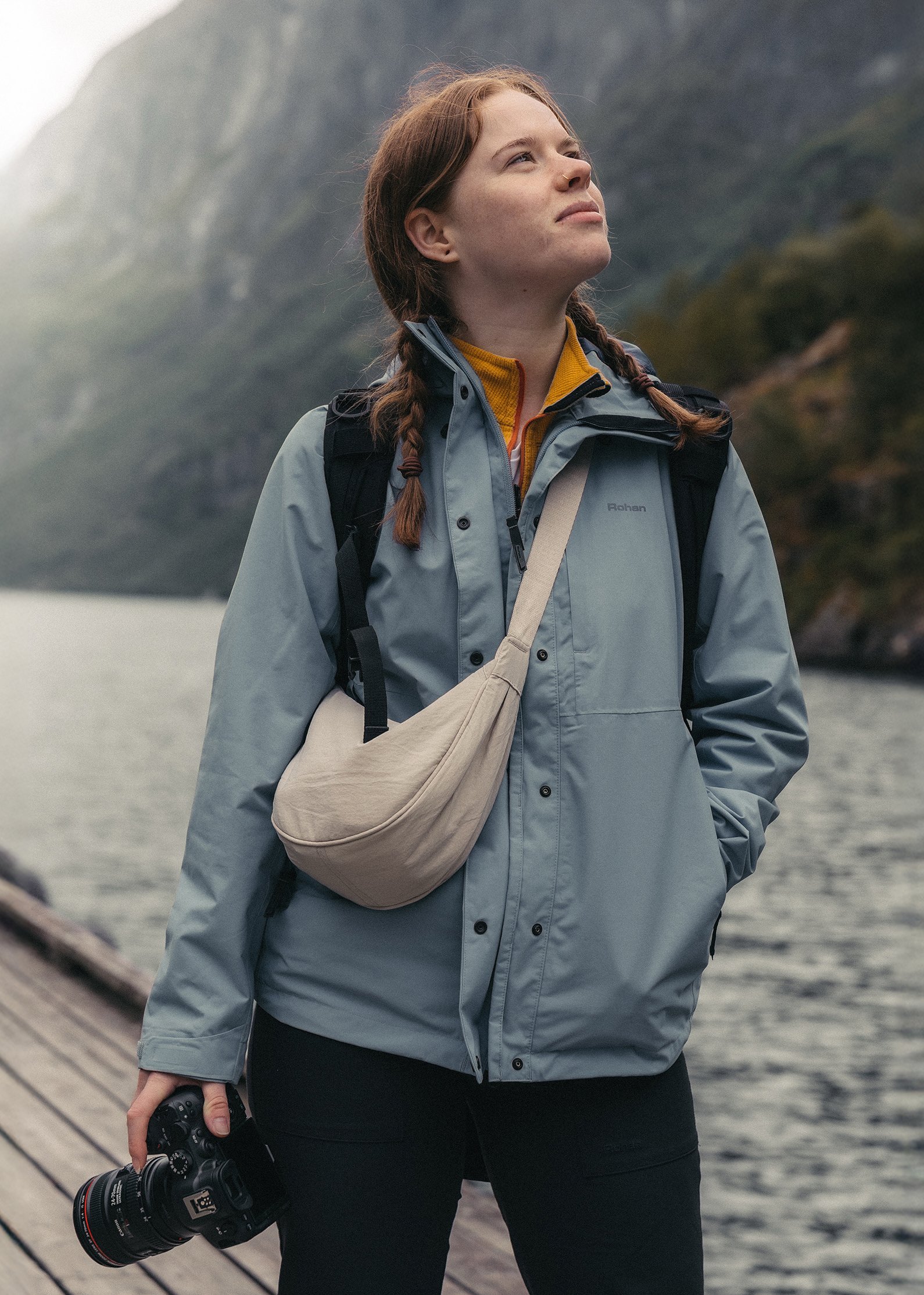 A woman with red hair in braids standing outdoors near water, holding a camera in her right hand, wearing a gray jacket, yellow shirt, backpack, and beige waist bag, looking up with mountains in the background.