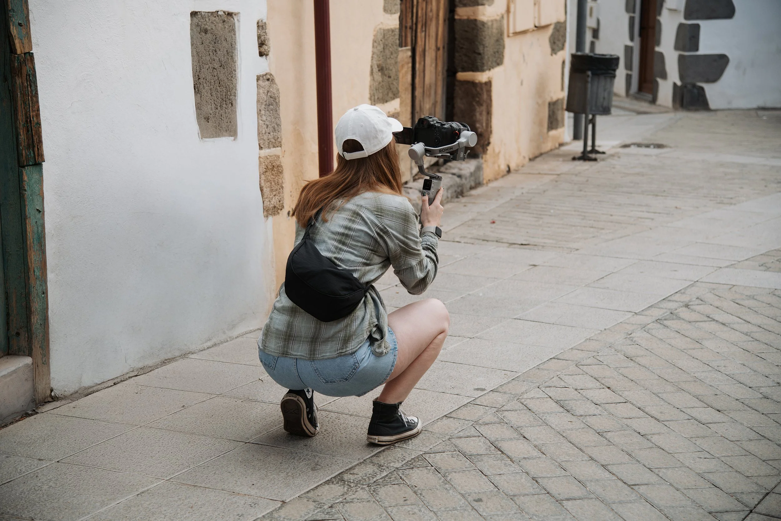A woman with long red hair, wearing a white cap, plaid shirt, denim shorts, and black sneakers, crouching on a sidewalk and holding a camera mounted on a gimbal.
