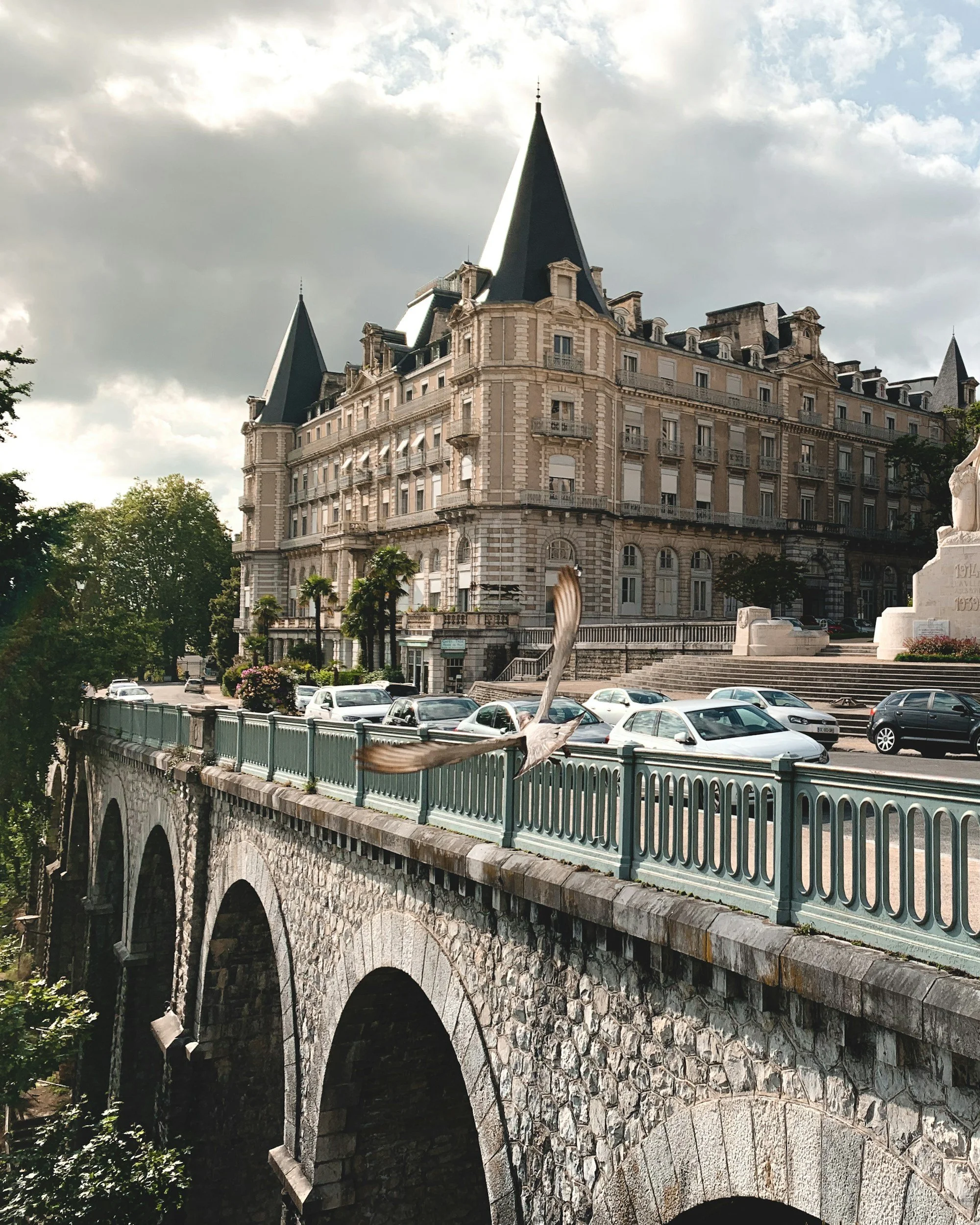 Un grand bâtiment de style château avec des tours pointues, situé au-dessus d'un pont en pierre avec une balustrade verte, et des voitures garées en dessous. Il y a aussi une statue blanche à droite, avec un ciel partiellement nuageux au-dessus.