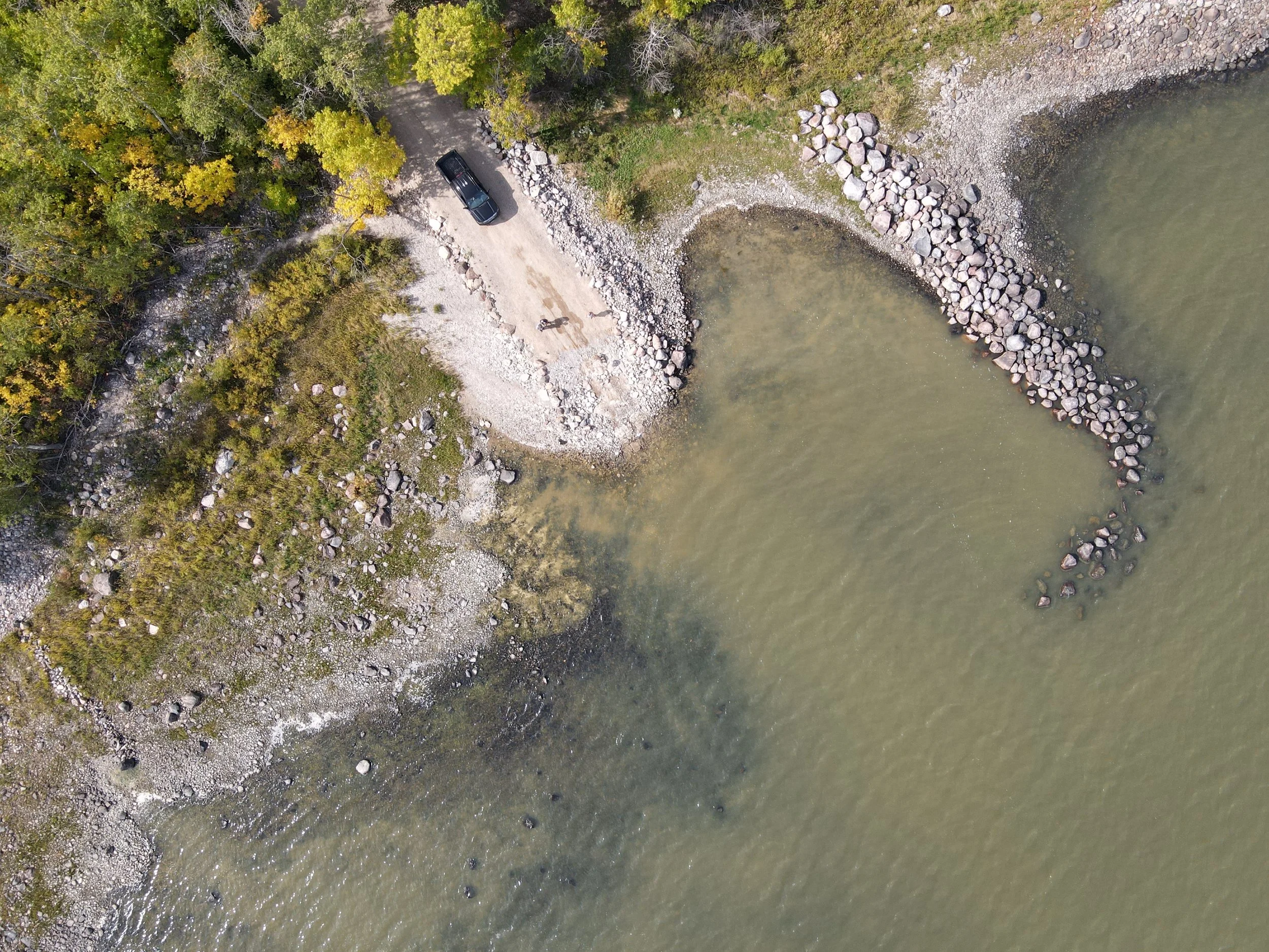 An aerial view of a lakeshore with a rocky breakwater curved into the water, a dirt road, a black vehicle, and a forested area with trees.