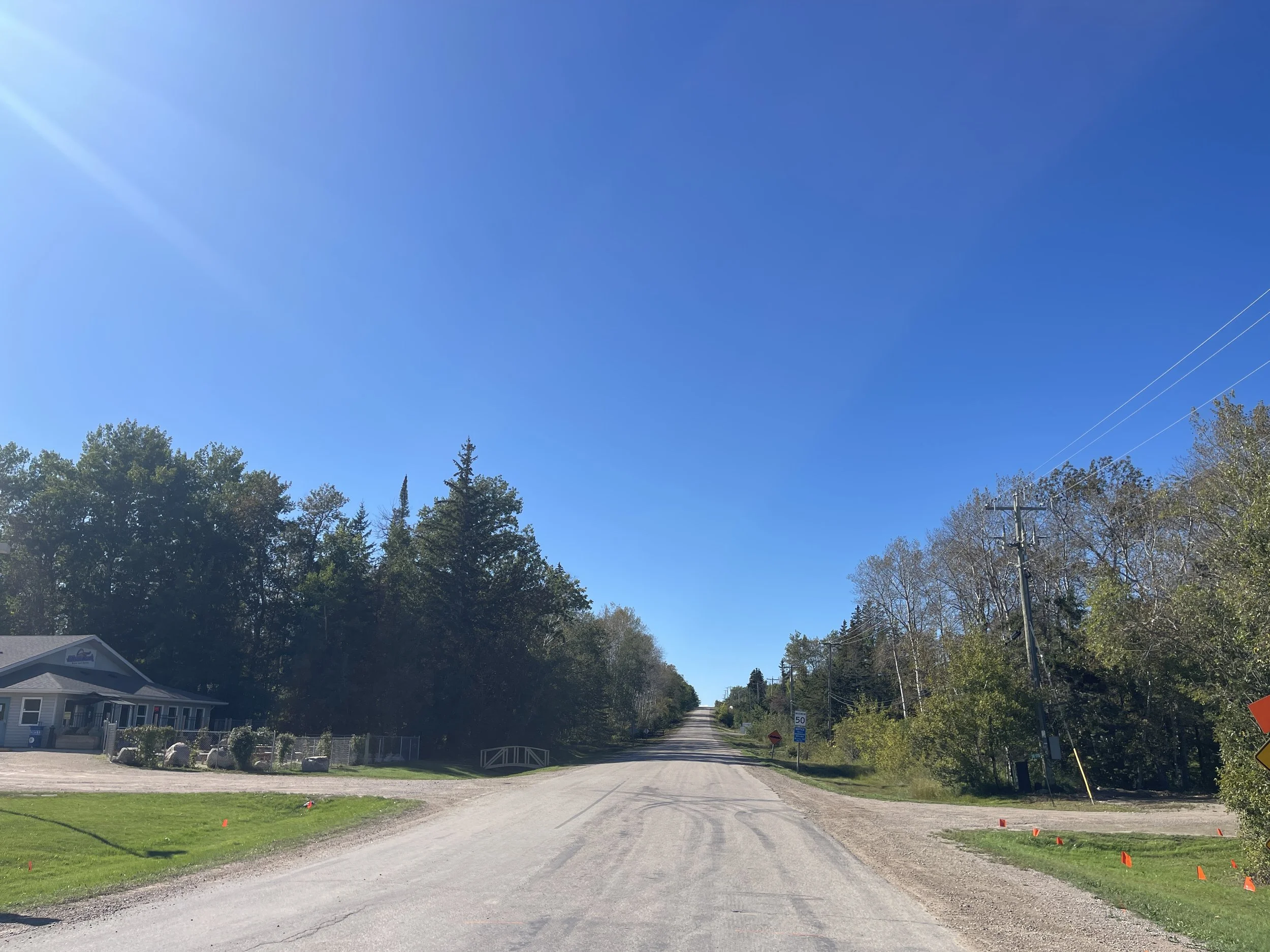 A rural road with a slight incline, surrounded by trees and clear skies, with small orange flags on both sides and utility poles on the right.