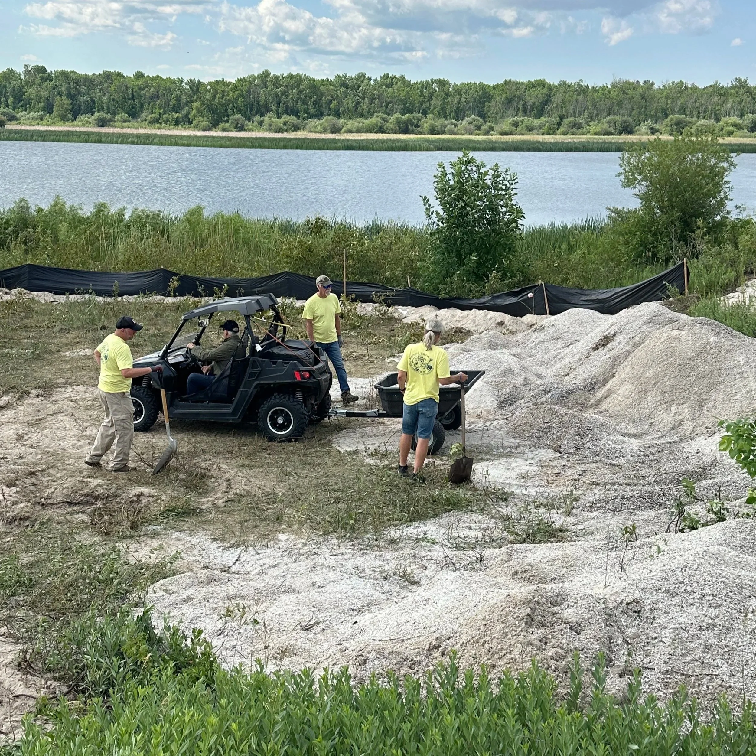 People working outdoors near a body of water, with a small off-road vehicle and shovels, on a rocky, grassy area surrounded by trees.